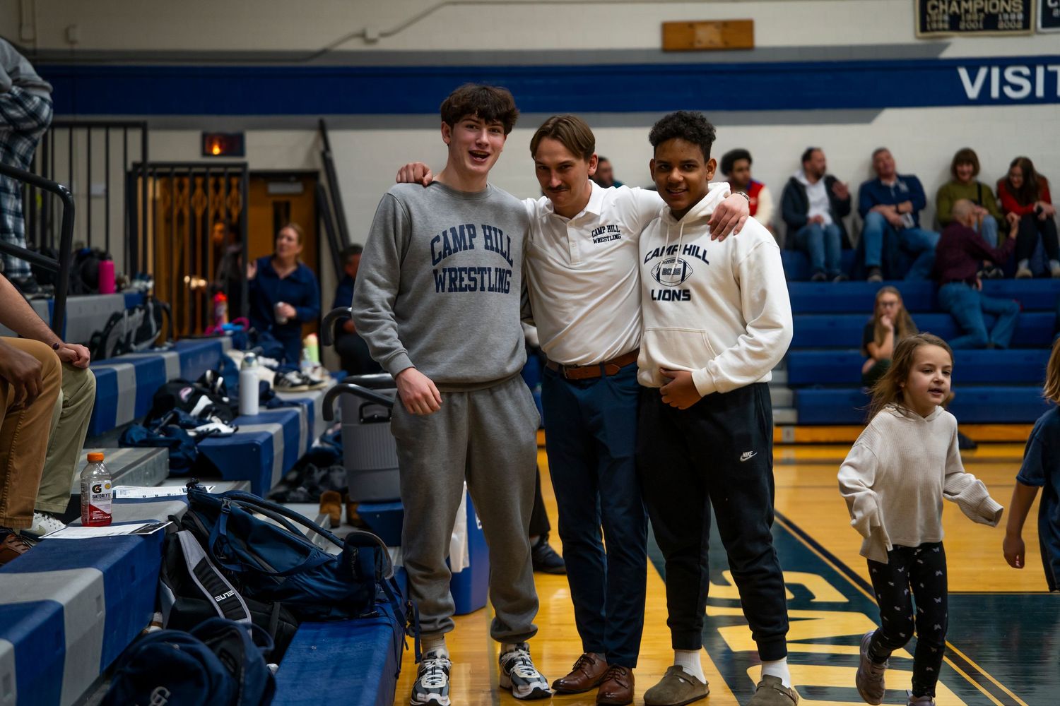Three smiling wrestlers pose together courtside at an indoor high school wrestling tournament.