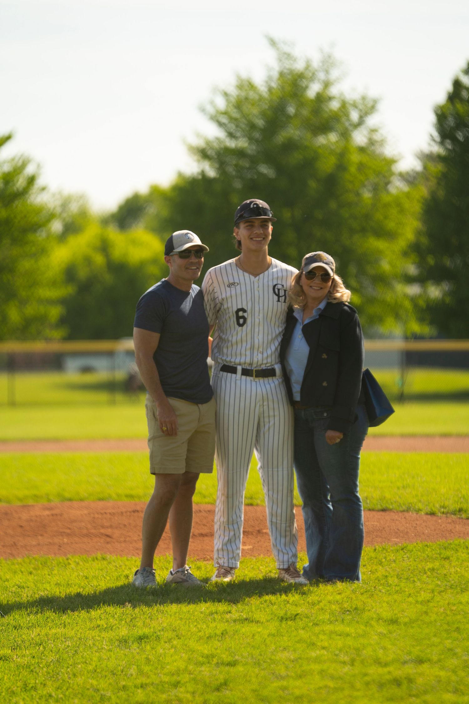 Baseball player #6 in pinstripe uniform poses with two smiling parents on a sunny green baseball field.
