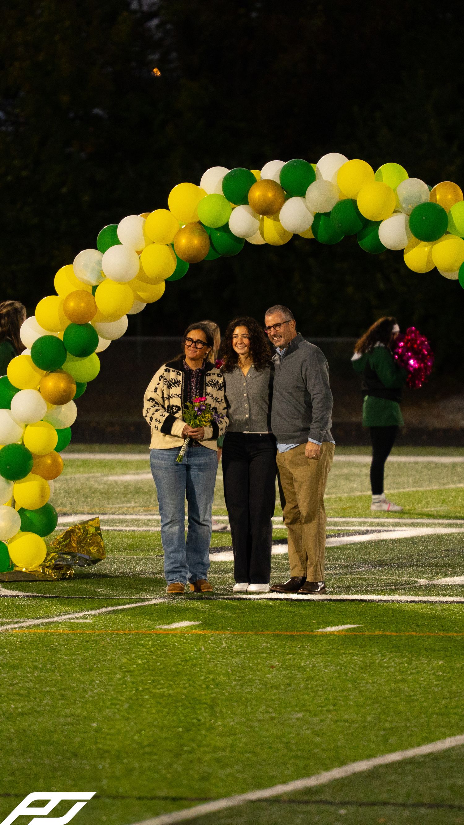Three people pose under a green, yellow, white, and gold balloon arch on a football field at a school ceremony.