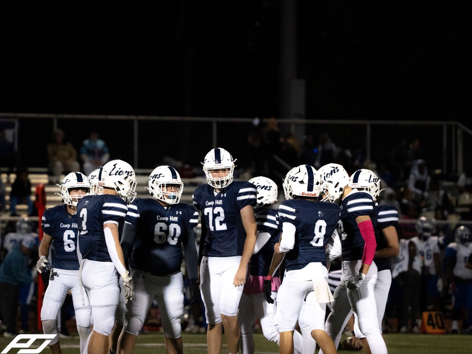 High school football team in navy uniforms huddles on field during night game under stadium lights.