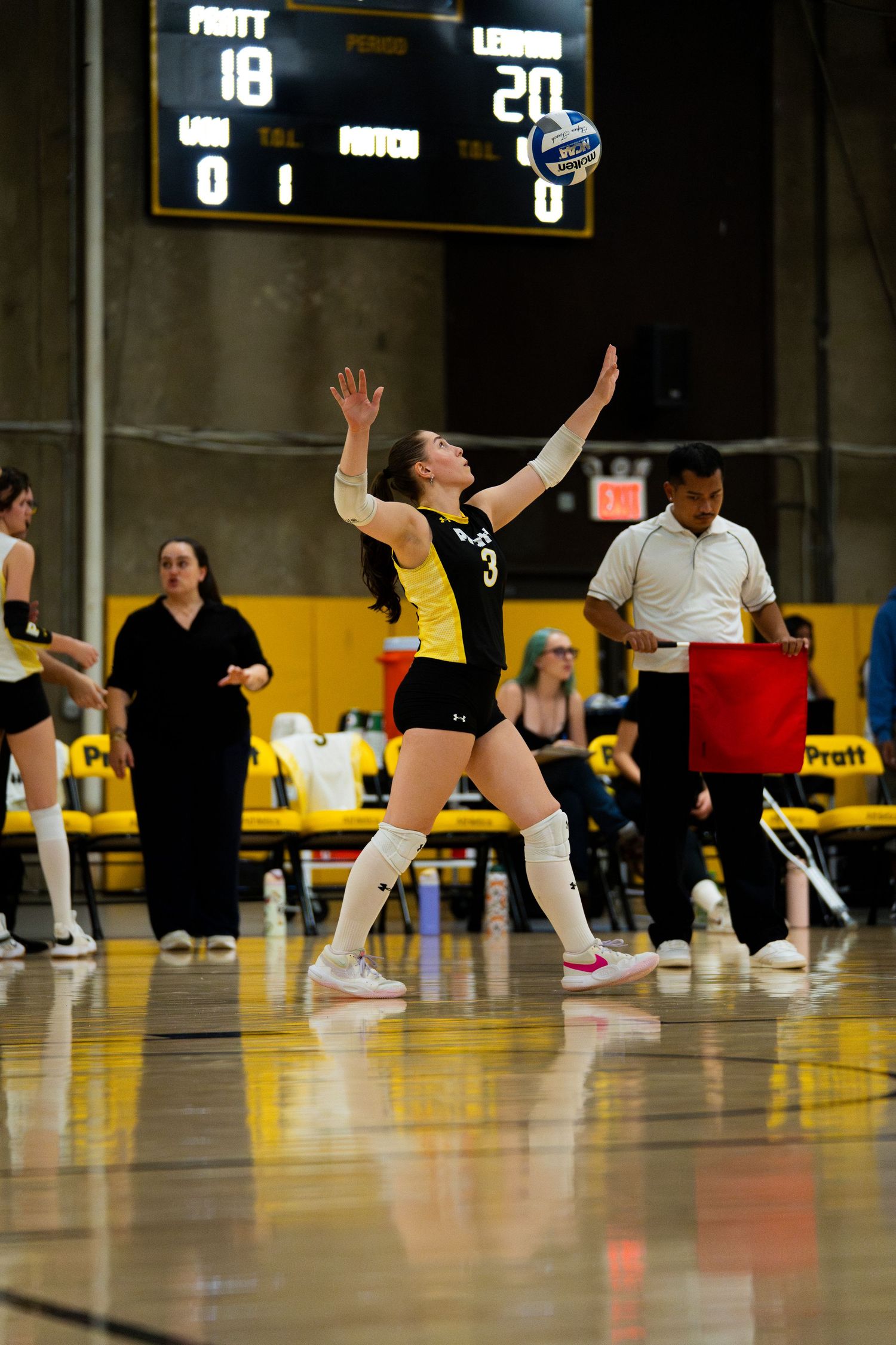 Volleyball player in black and gold uniform serves the ball during a match with scoreboard showing Pratt vs opponent.