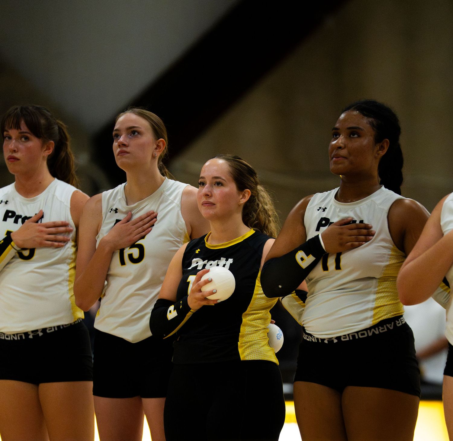 Volleyball players in black and gold uniforms stand with hands on hearts during national anthem ceremony.