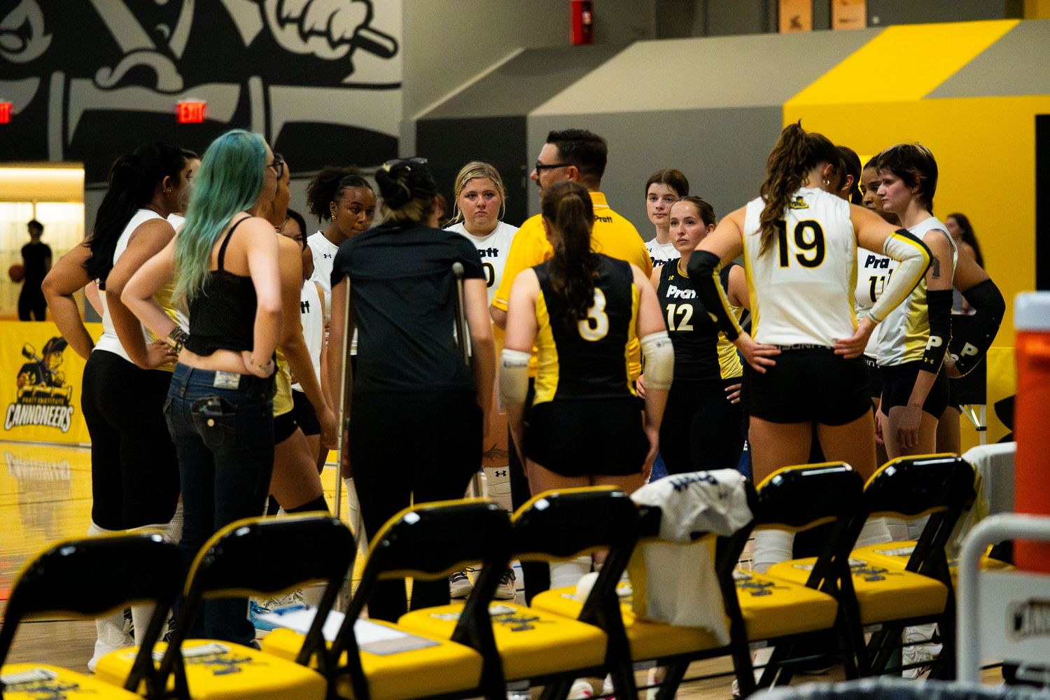 Volleyball team huddles during timeout in black and gold uniforms on indoor court.