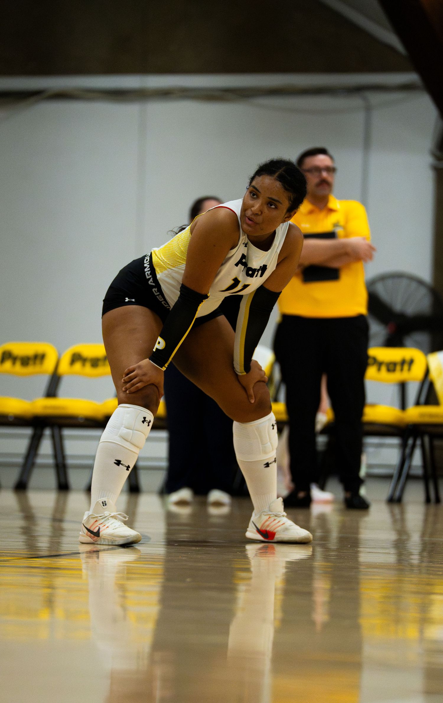Female volleyball player in black and white Pratt uniform crouches in ready position on indoor court during match.