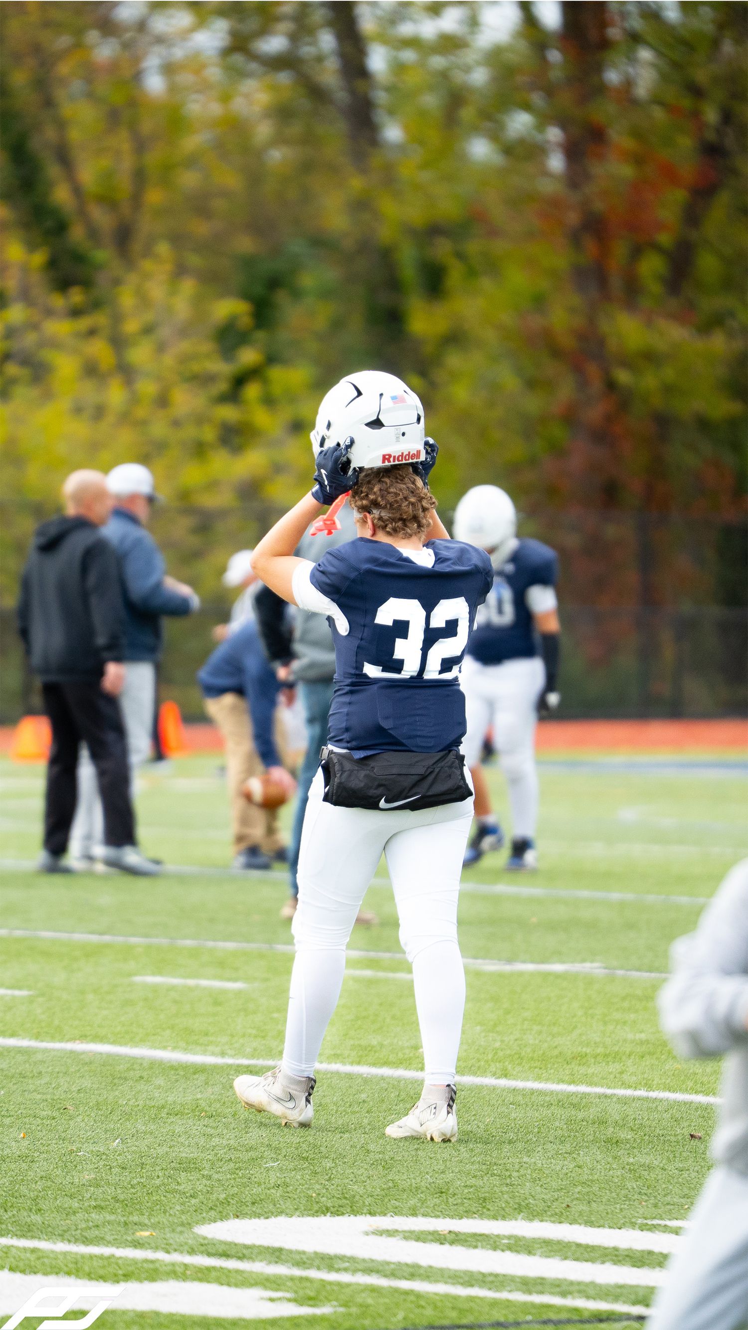 Football player number 32 in navy uniform carries helmet on autumn field.