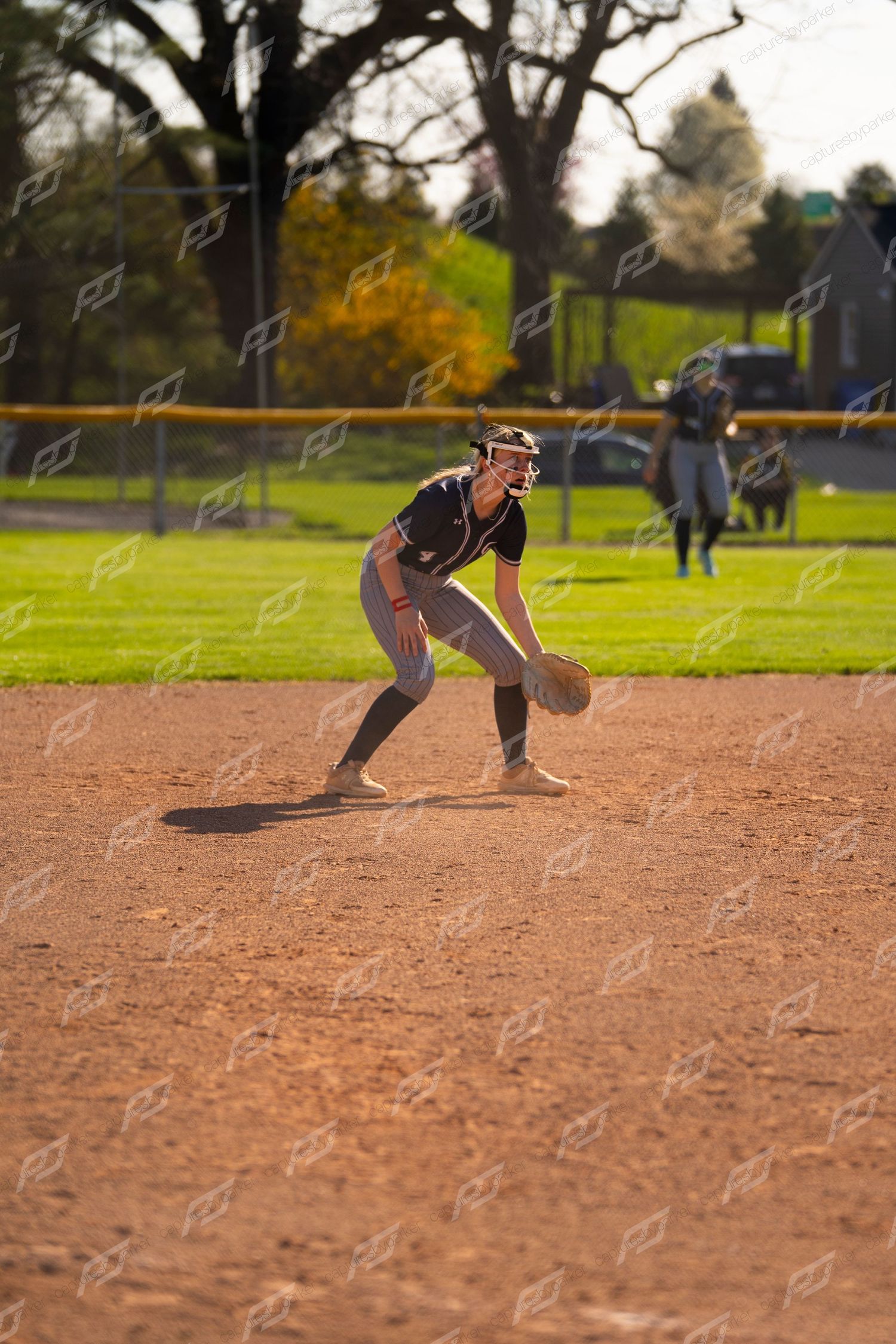 Female softball infielder in black uniform crouches in ready position on the dirt infield.