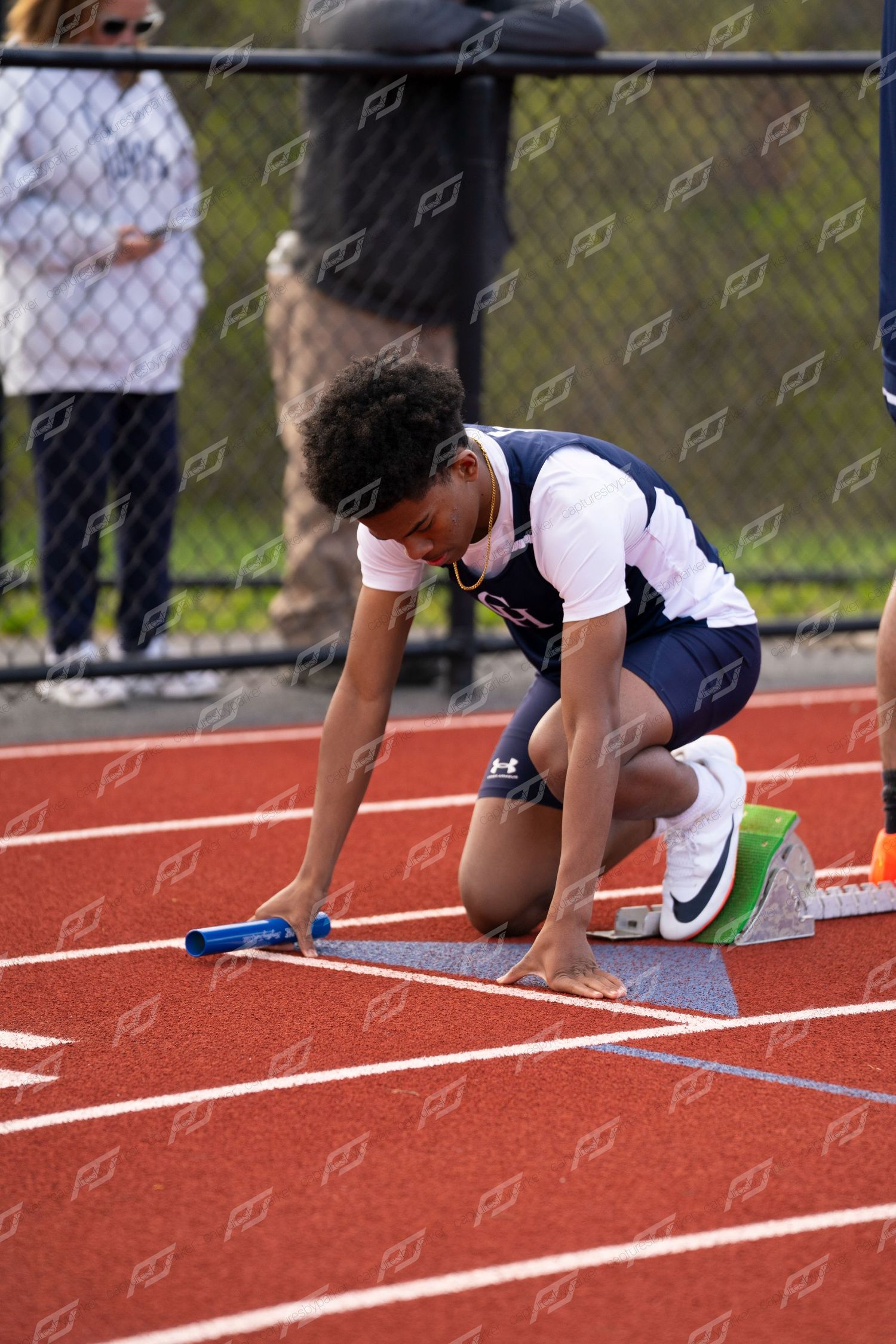 Male high school sprinter in starting position on red track gripping blue relay baton at starting blocks.
