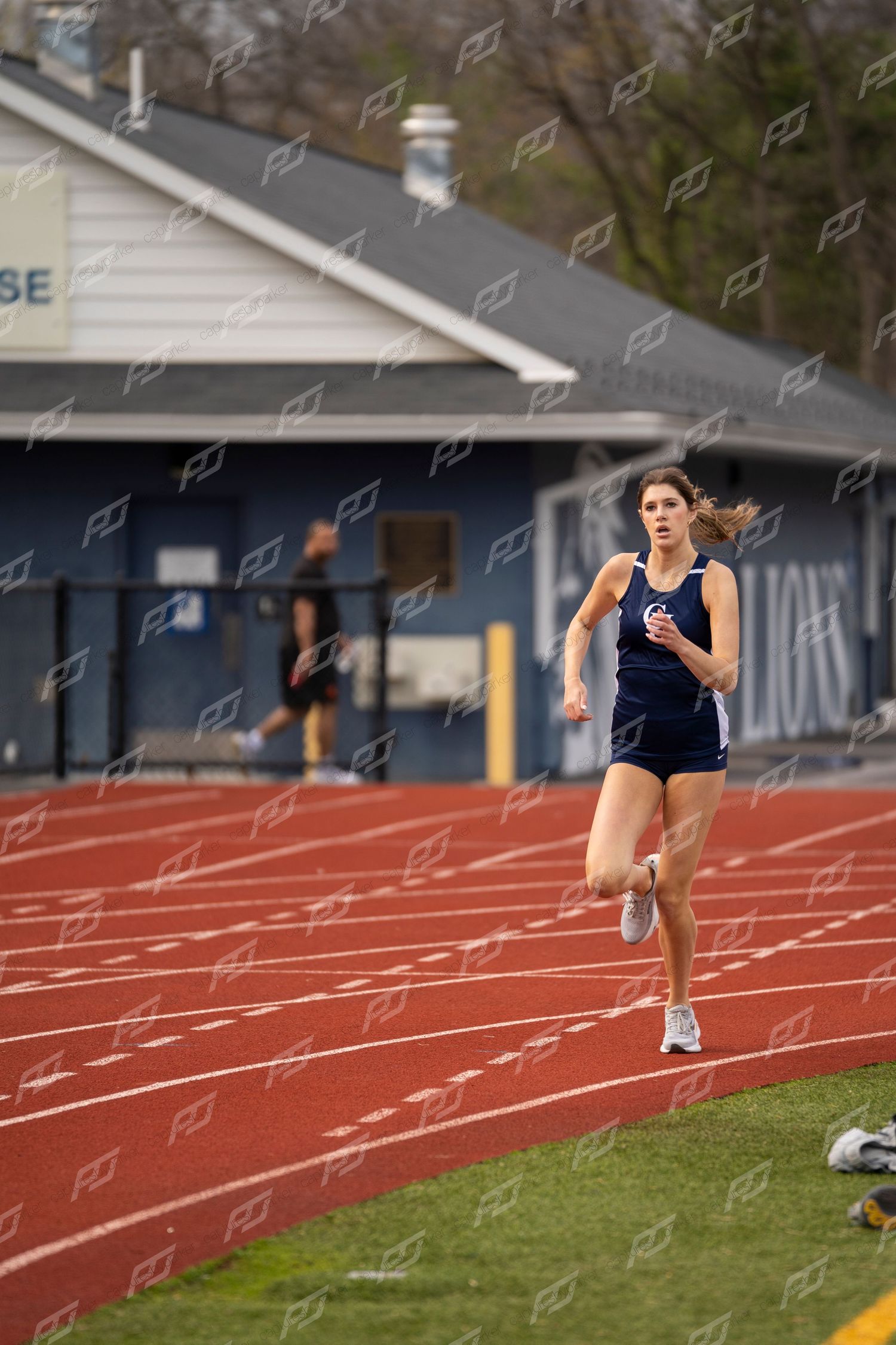 Female athlete in navy Lions uniform runs alone on red track during outdoor meet.