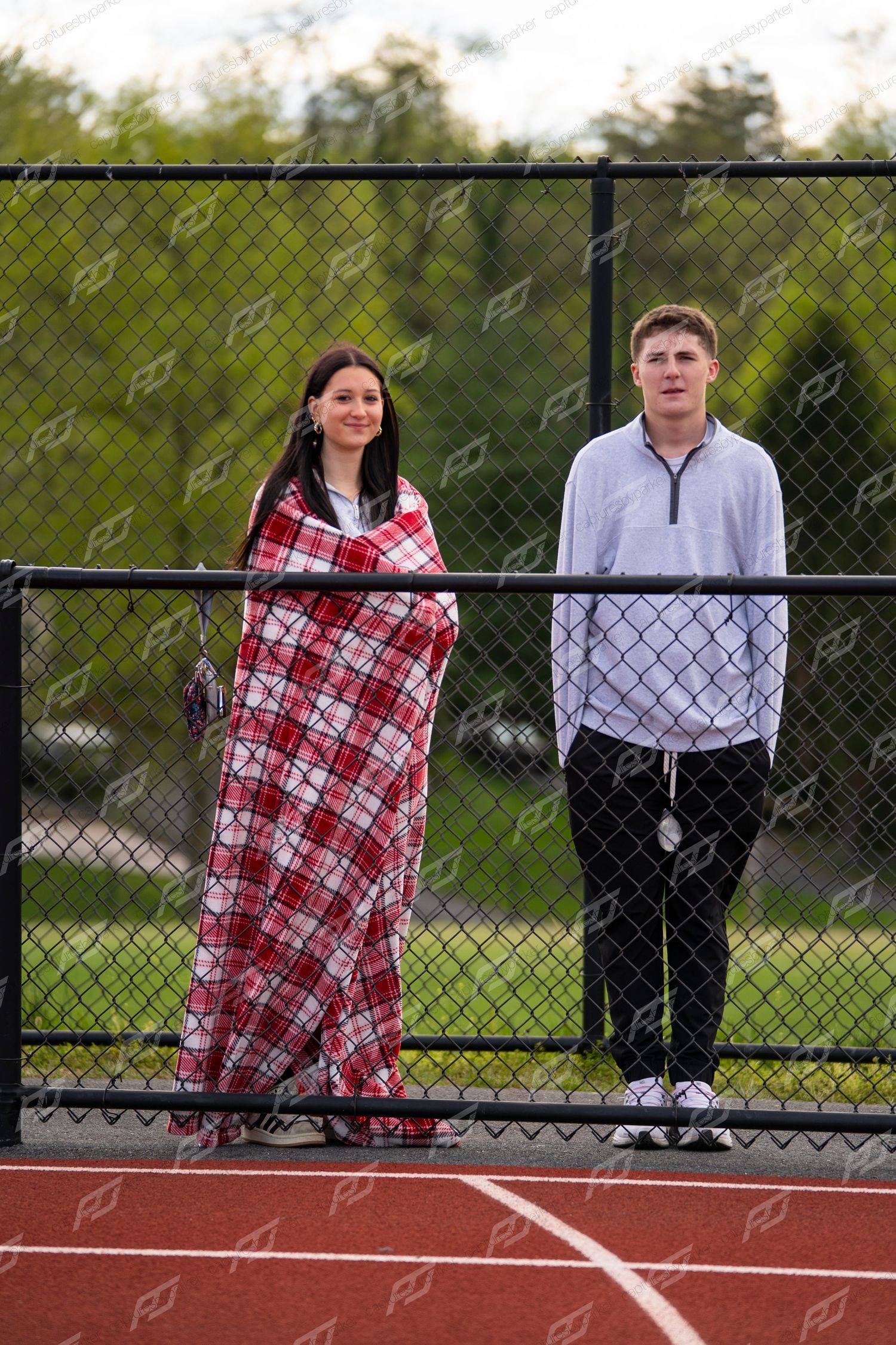 Two young spectators standing by chain-link fence at track meet, girl wrapped in red plaid blanket.