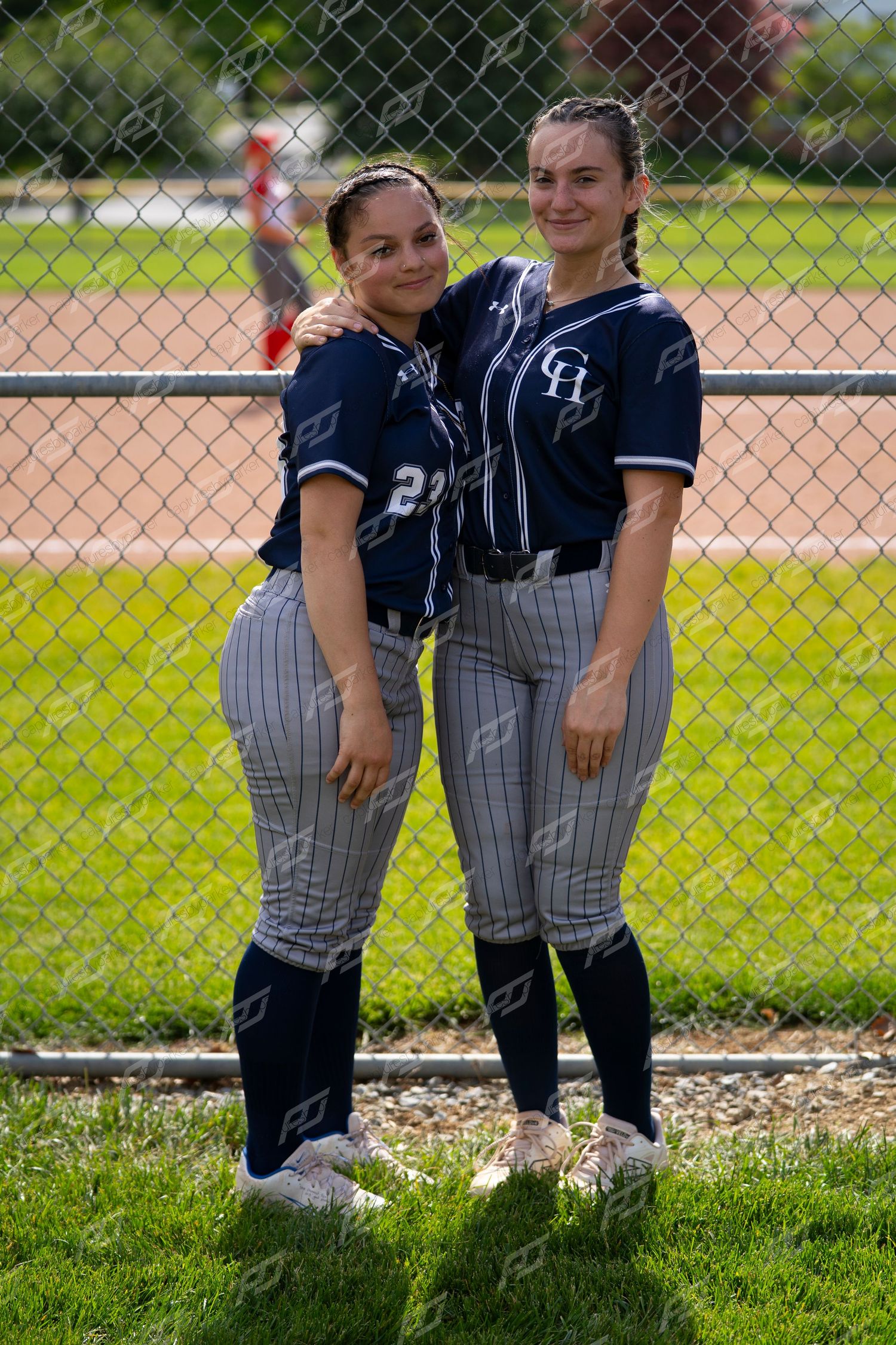 Two female softball players in navy GH uniforms smile and pose together by a fence.