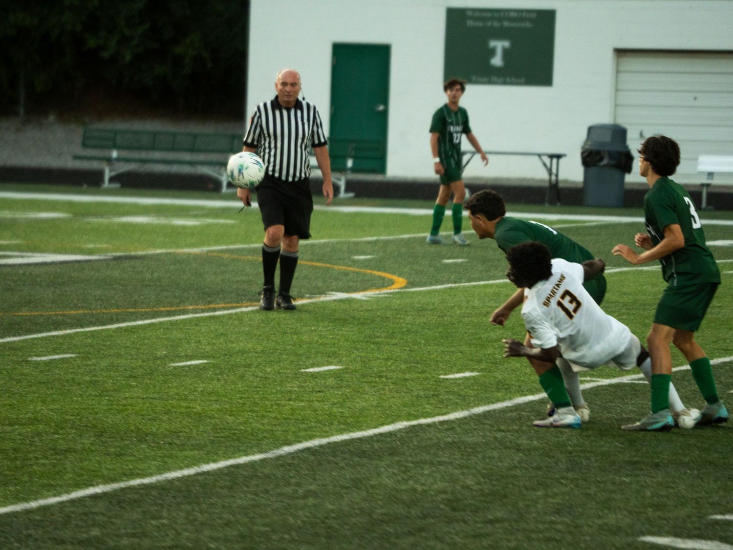 High school soccer player number 13 heads the ball near two defenders while a referee watches closely.