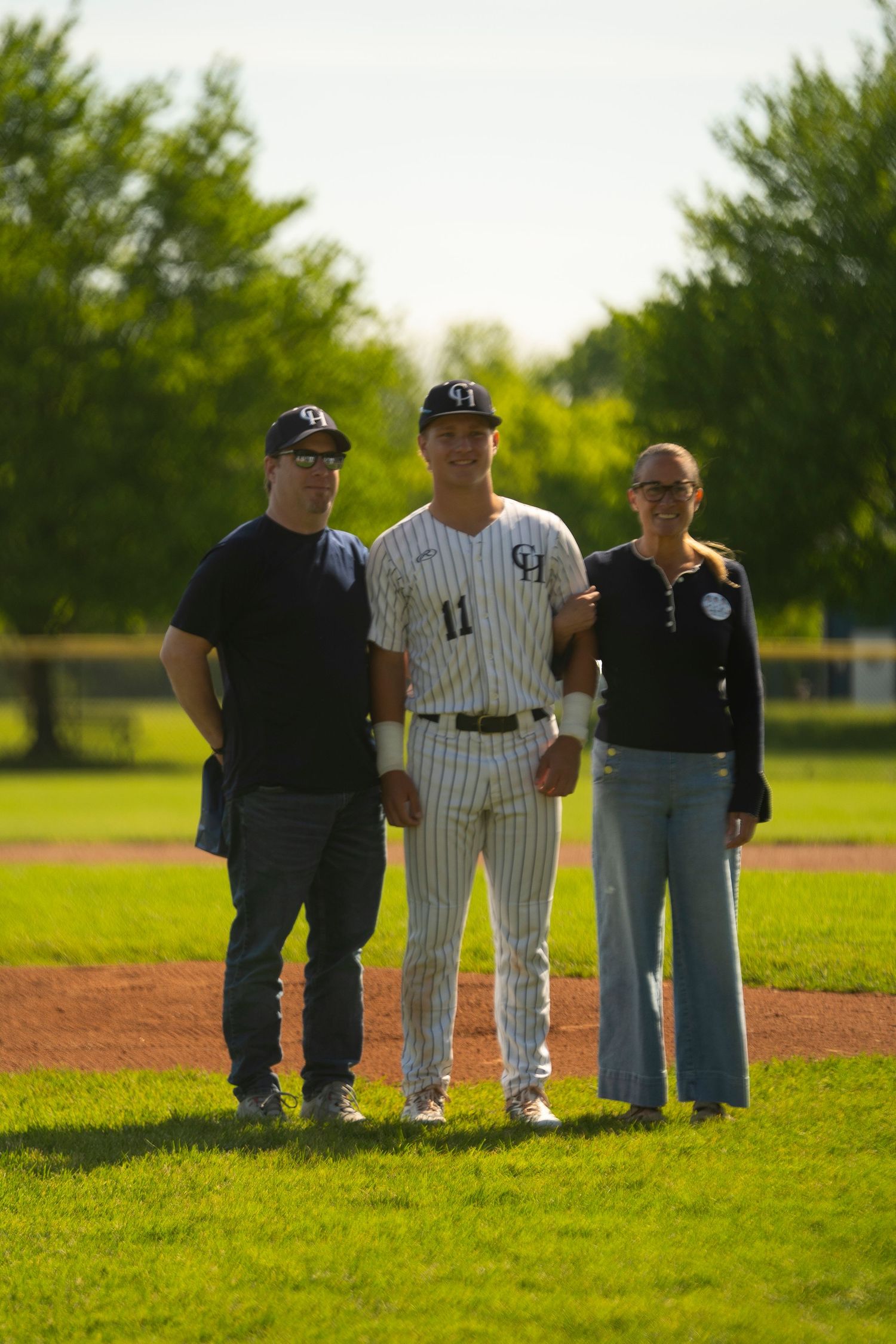 Baseball player #11 in pinstripe uniform poses with two proud parents on a sunny green baseball field.
