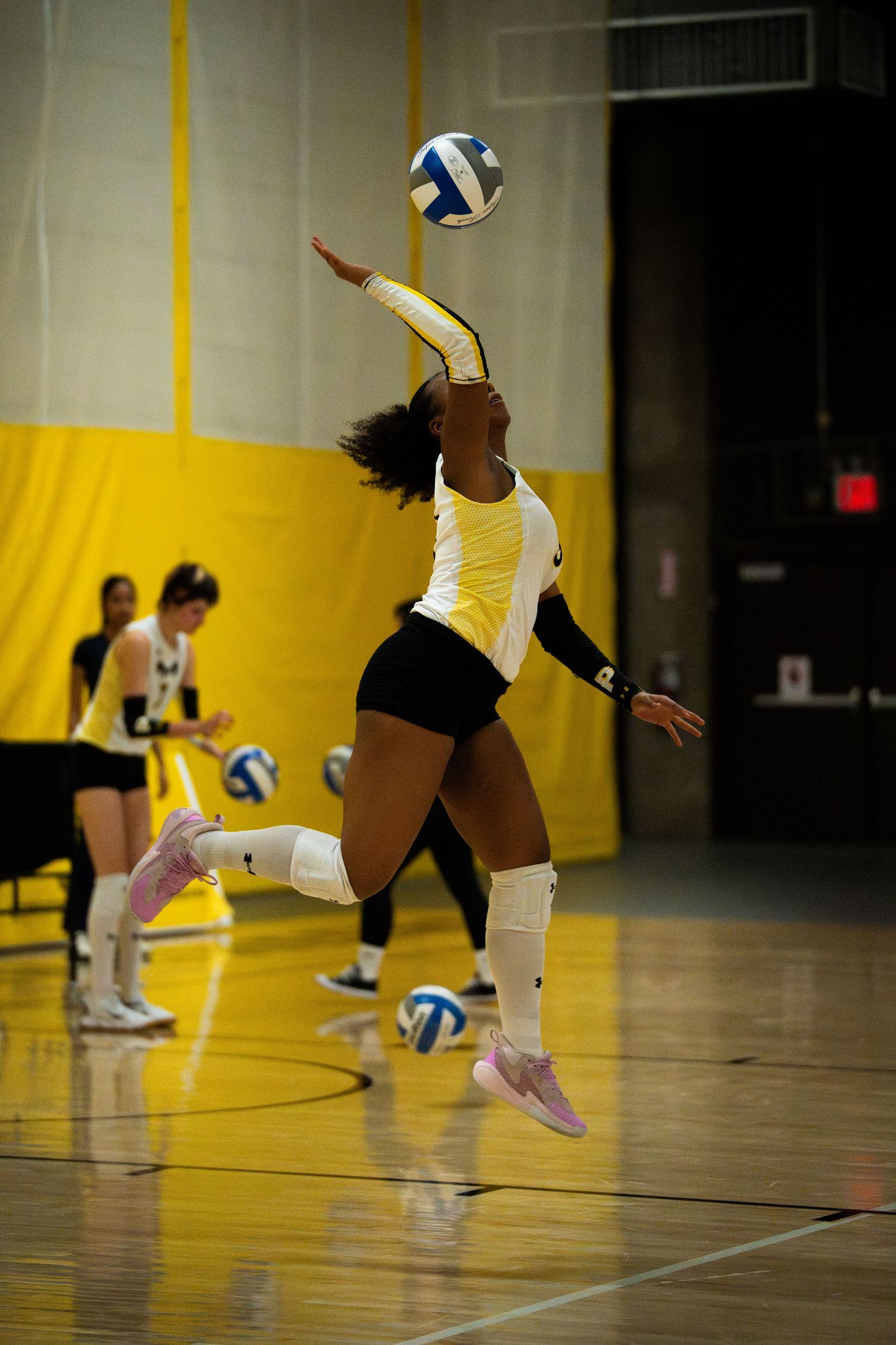Female volleyball player in yellow and white uniform jumping to spike the ball during an indoor match.