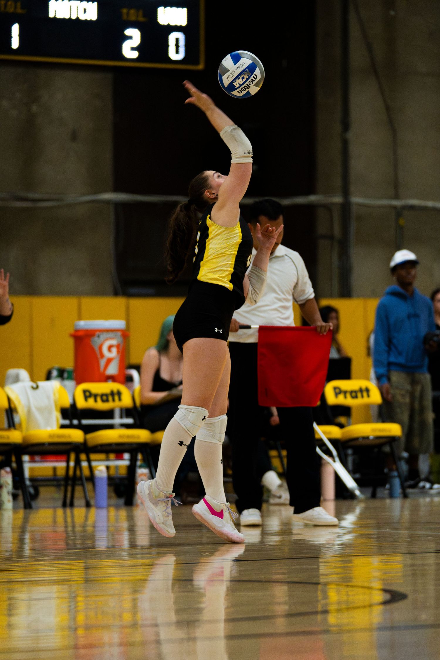 Female volleyball player in yellow uniform jumps to spike the ball during an indoor match with scoreboard visible.