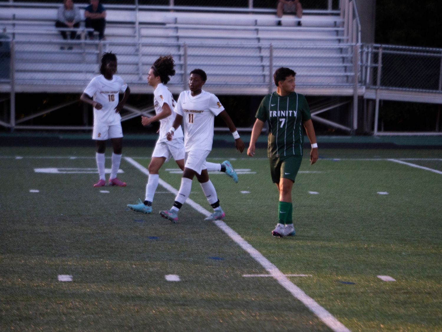 White-uniformed soccer players celebrate near Trinity player number 7 on a green turf field.