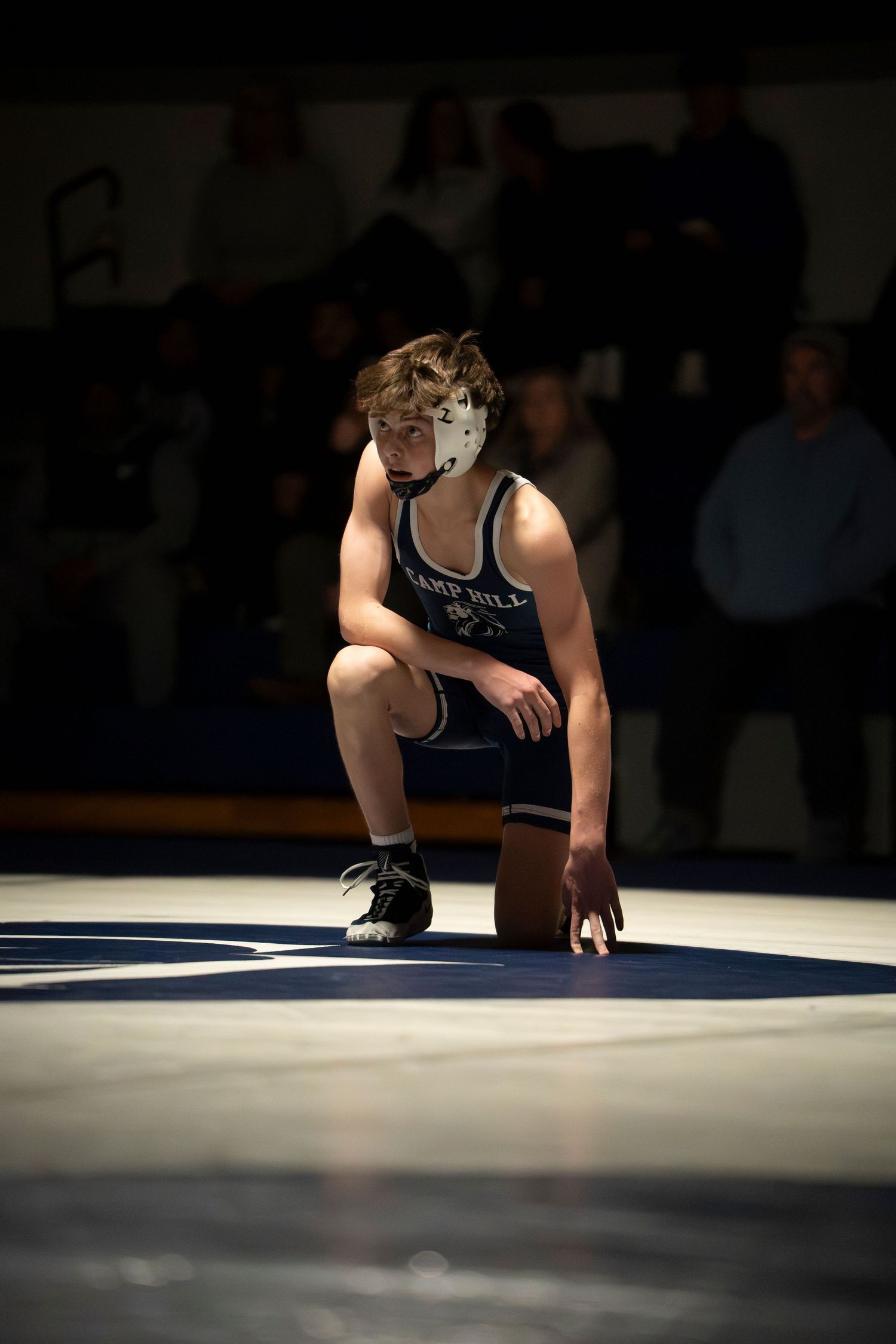 A Camp Hill wrestler in a navy singlet crouches in a ready stance on the wrestling mat during competition.