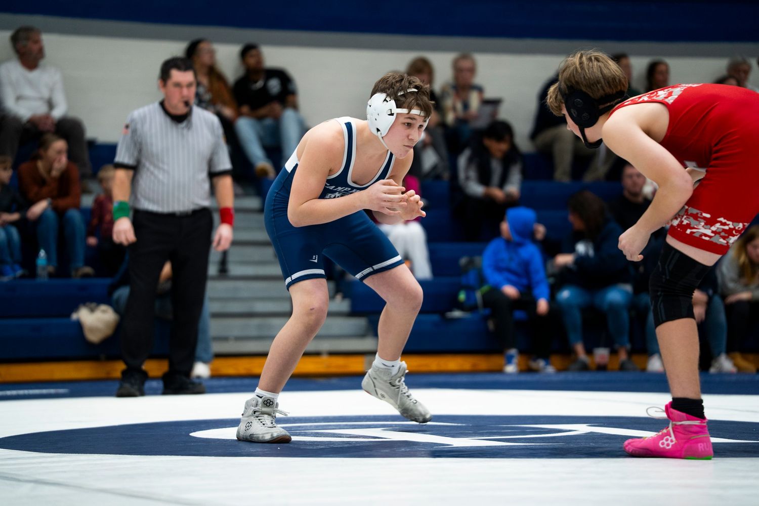 High school wrestler in blue singlet crouches in defensive stance facing red-uniformed opponent on mat.