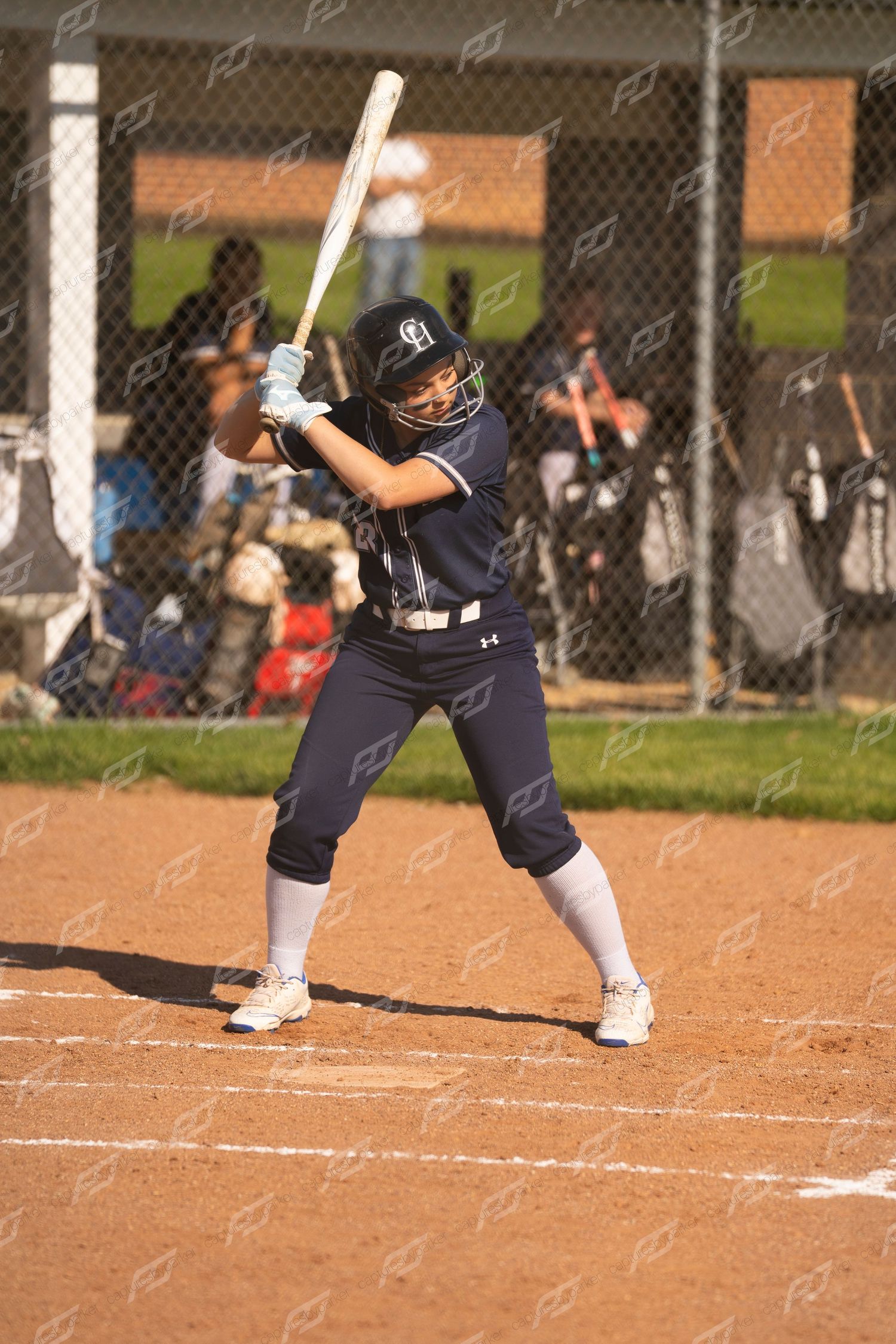 Female softball batter in navy uniform stands ready at home plate in batting stance.