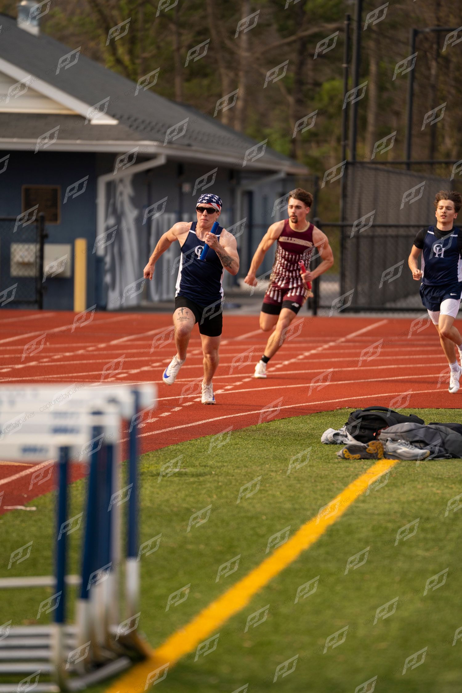 Three male runners compete in relay race on red track during outdoor track meet.