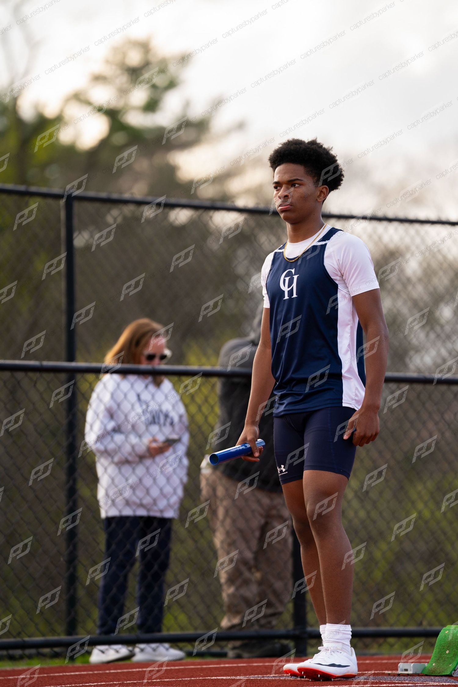 Young male track athlete in navy uniform holds blue baton while waiting at relay exchange zone during outdoor meet.