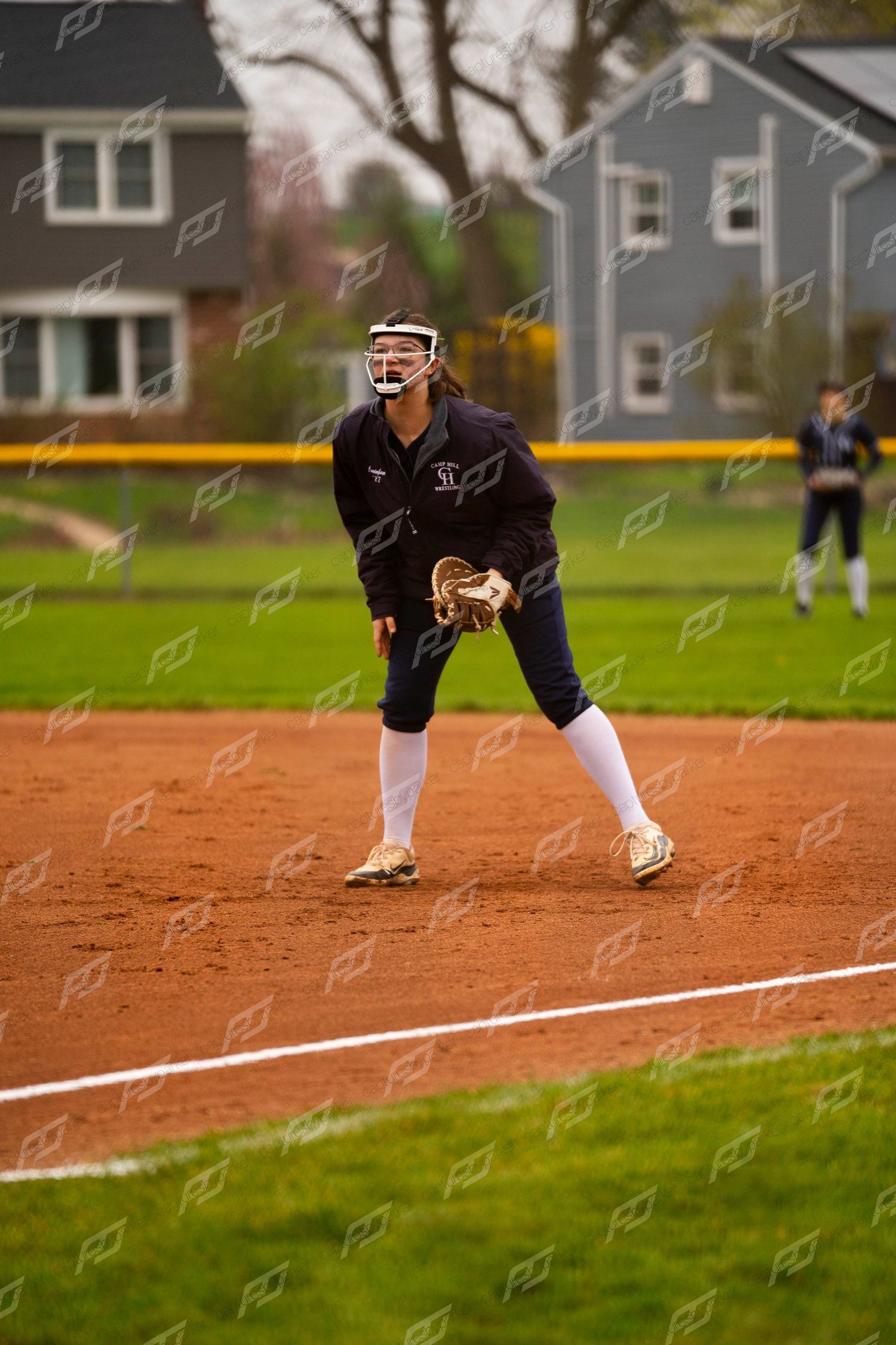 Softball player in dark uniform crouching in ready defensive position on infield dirt during a game.