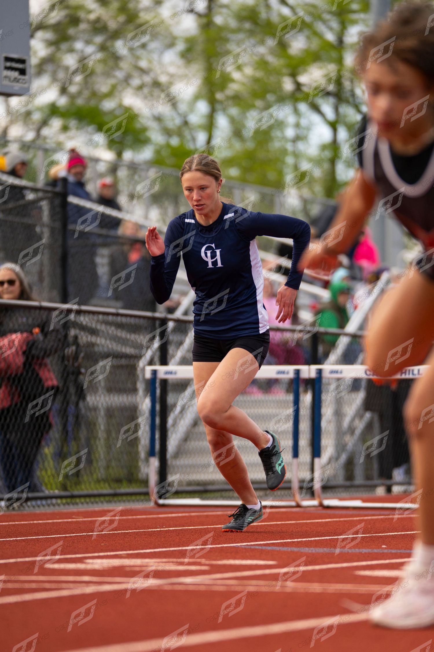 Female track athlete in navy CH uniform sprinting past hurdles on red track at outdoor meet.