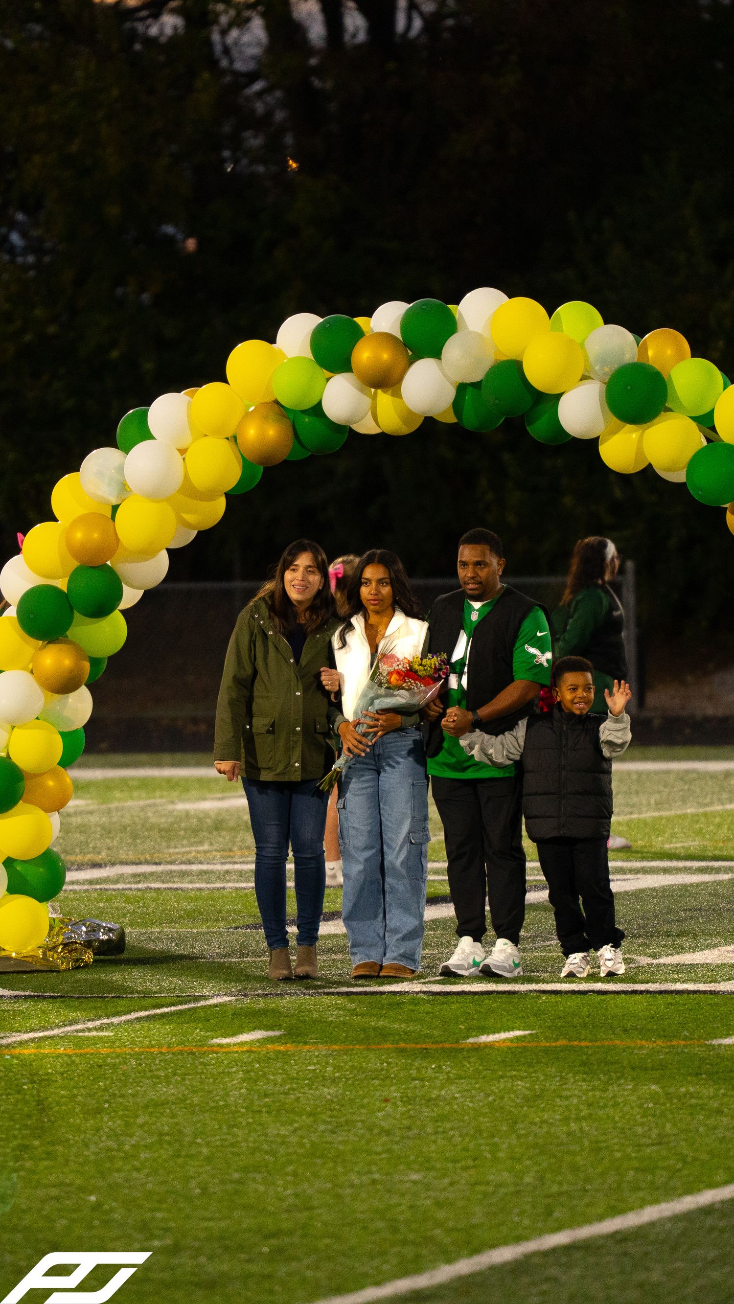 Senior athlete honored with flowers and family under green, gold, white balloon arch at evening field ceremony.