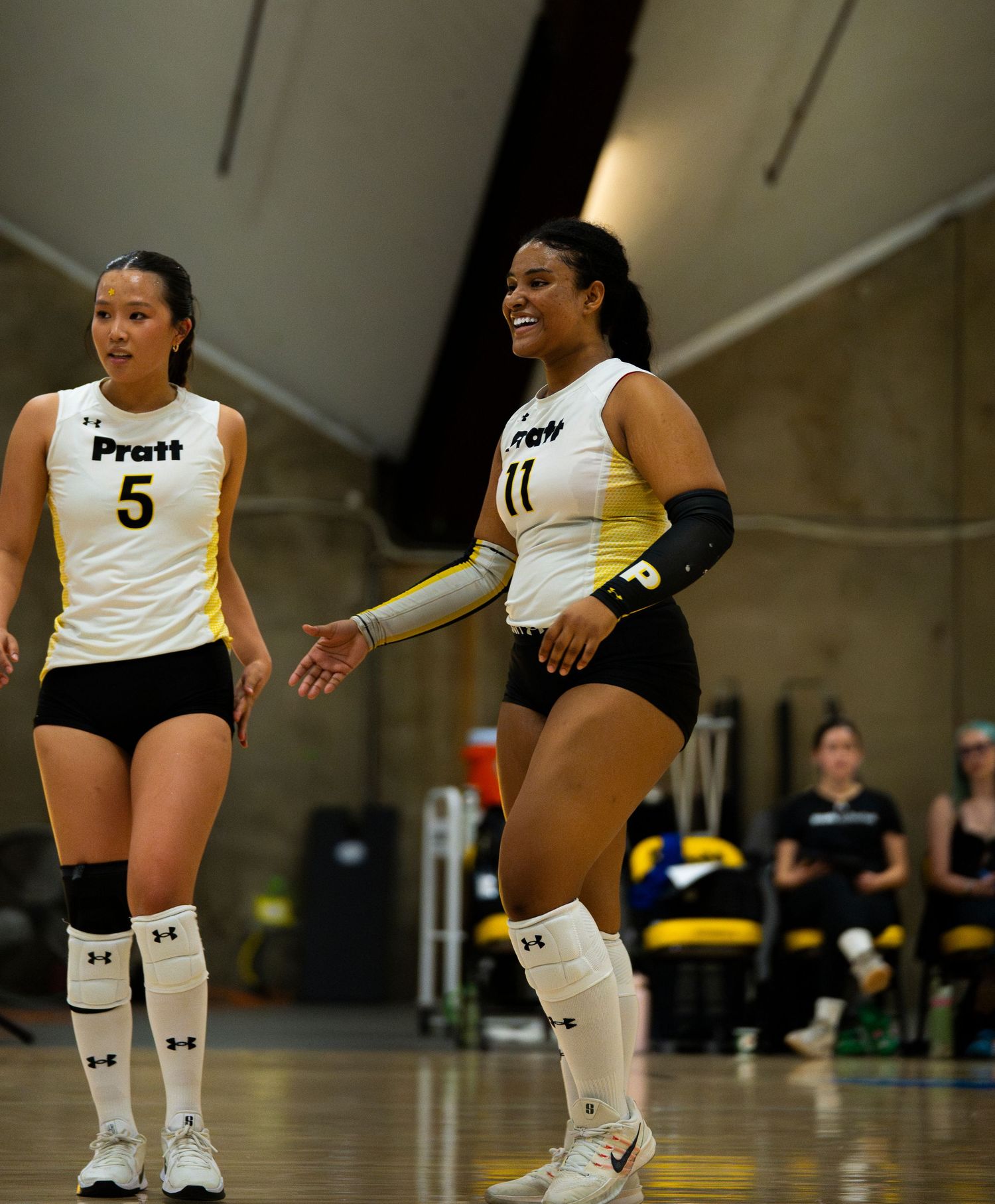 Two Pratt volleyball players in white and black uniforms smile during a match on an indoor court.