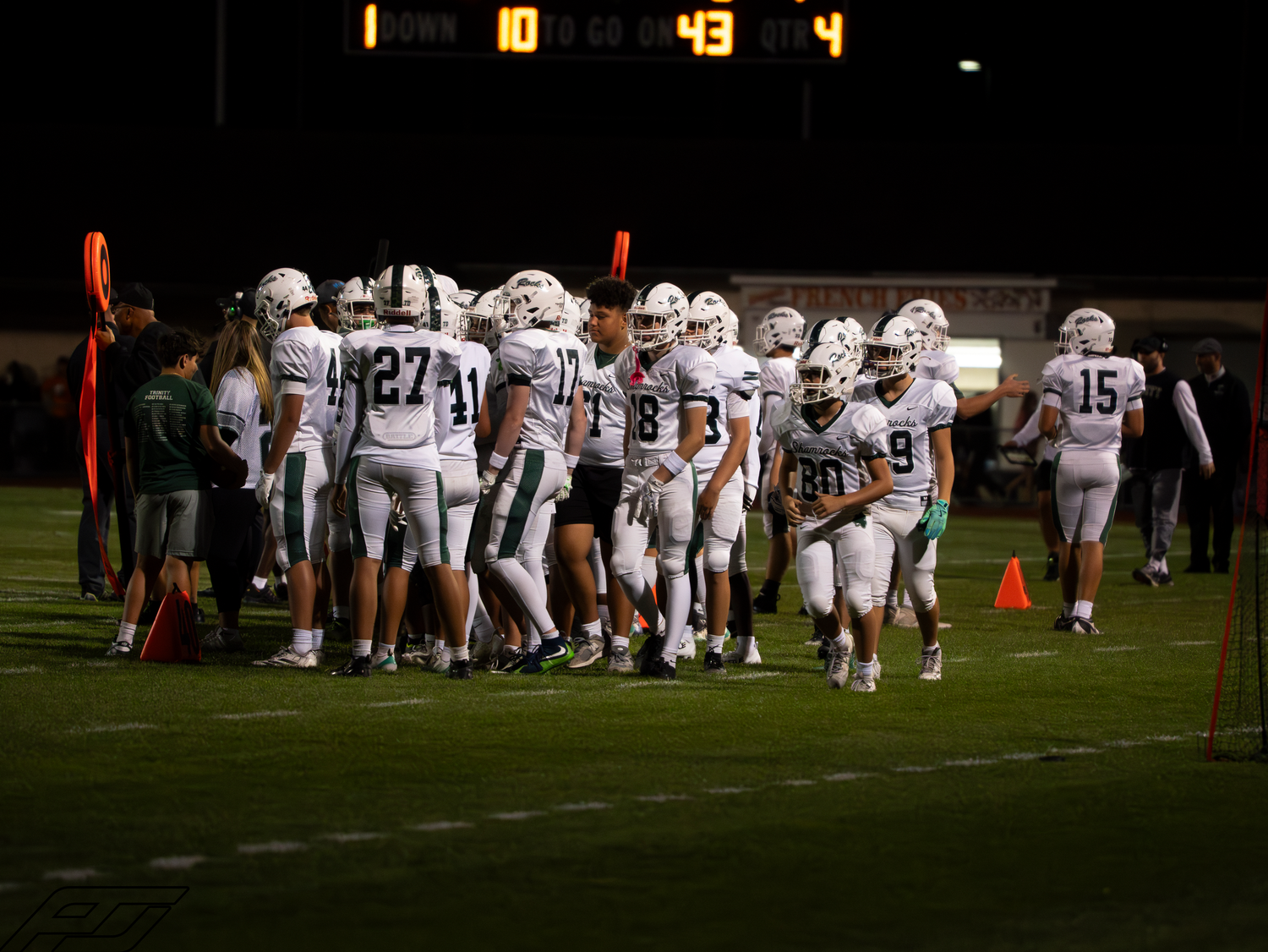 High school football team huddles on the sideline with a scoreboard showing 10-43 during a night game.