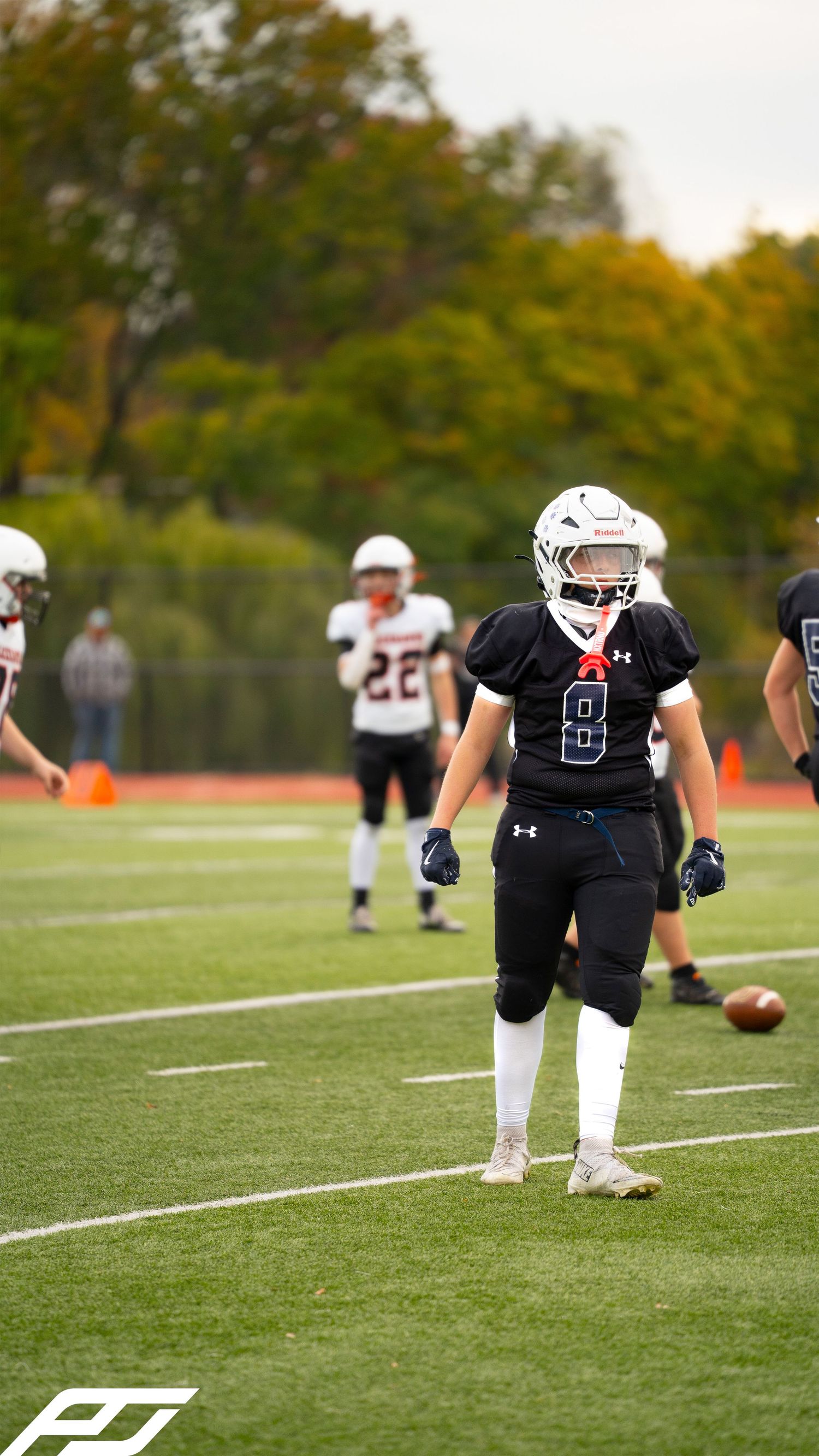 Youth football player number 8 in black Under Armour uniform stands on green turf field during an autumn game.