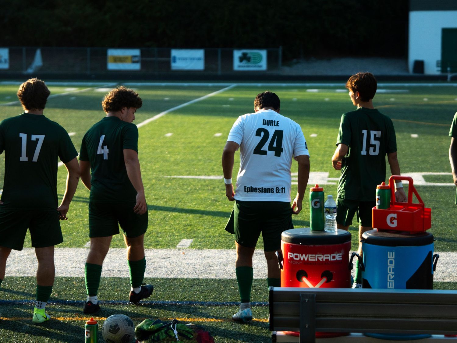 High school soccer players wearing green and white uniforms stand along the sideline during a game.