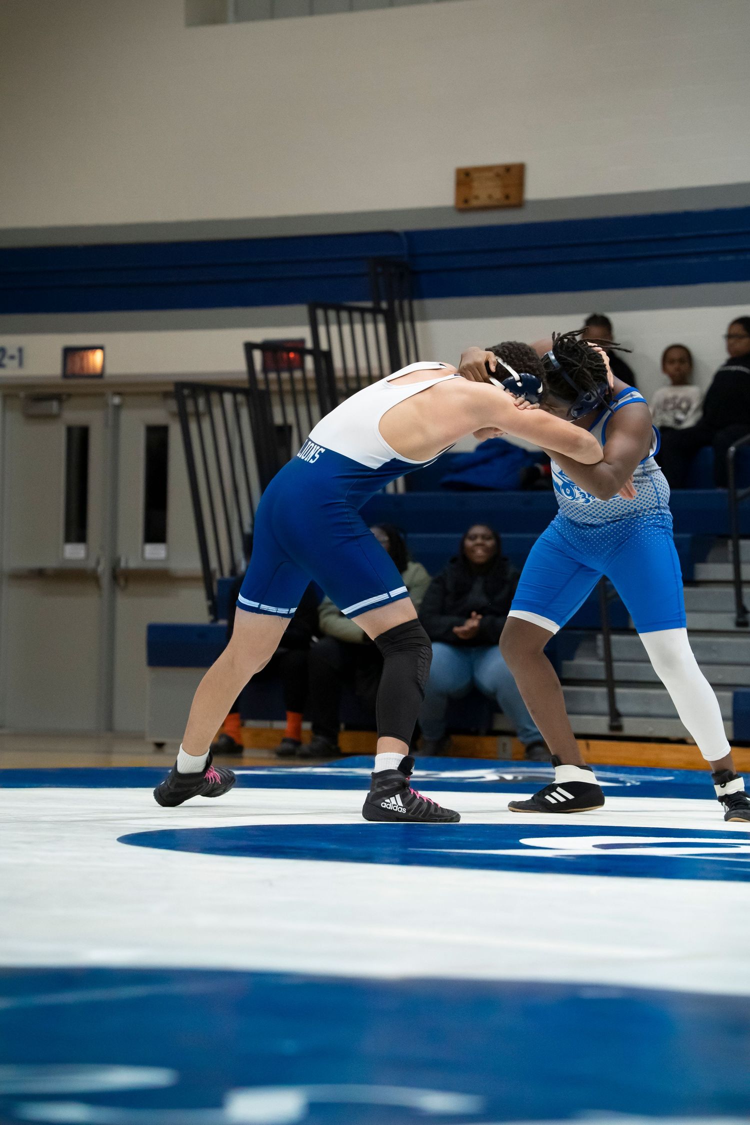 Two male wrestlers in blue and white singlets face off in a standing position during a high school wrestling match.