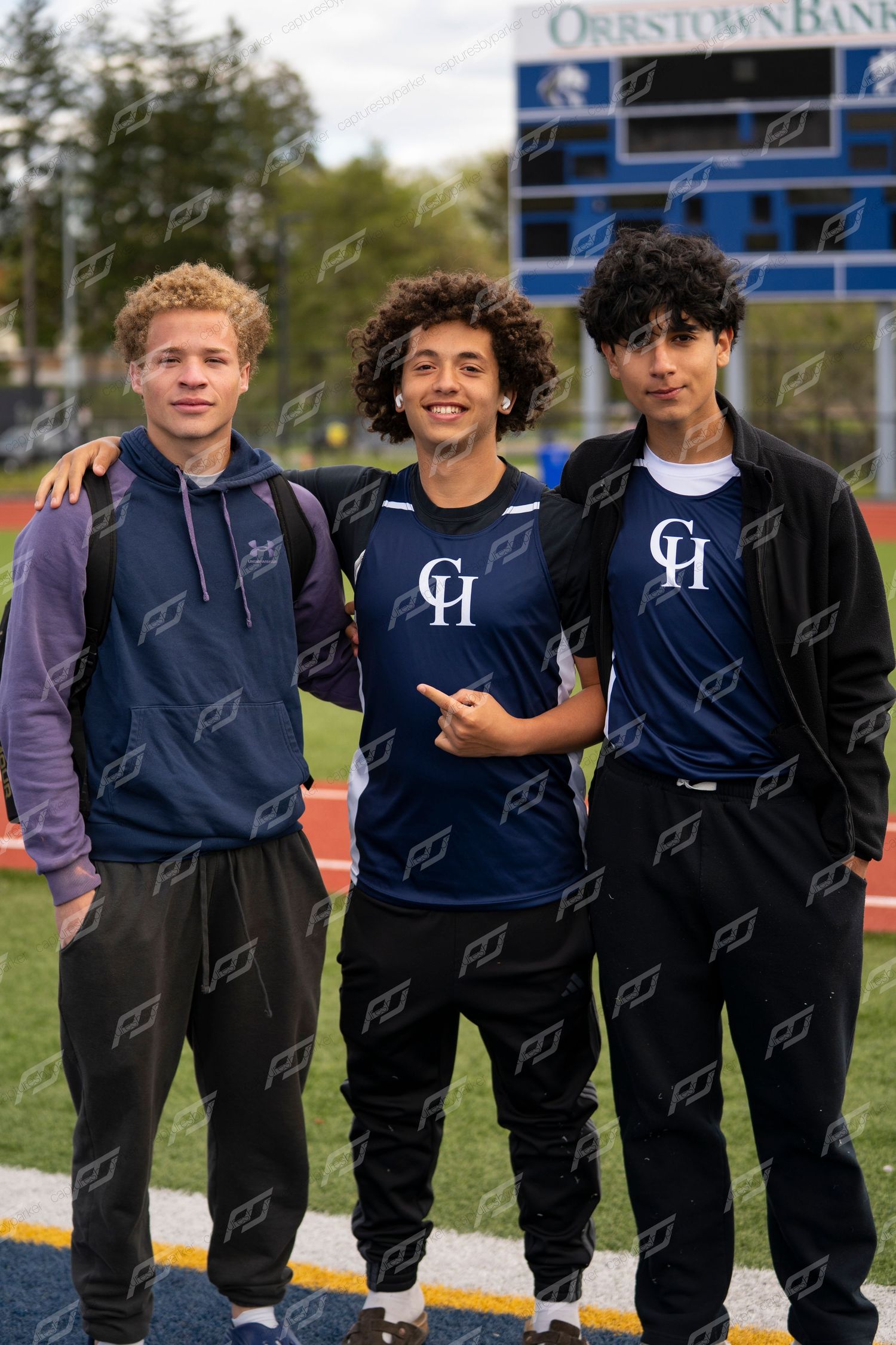 Three male CH track and field athletes posing together near an Orrstown Bank scoreboard at an outdoor meet.