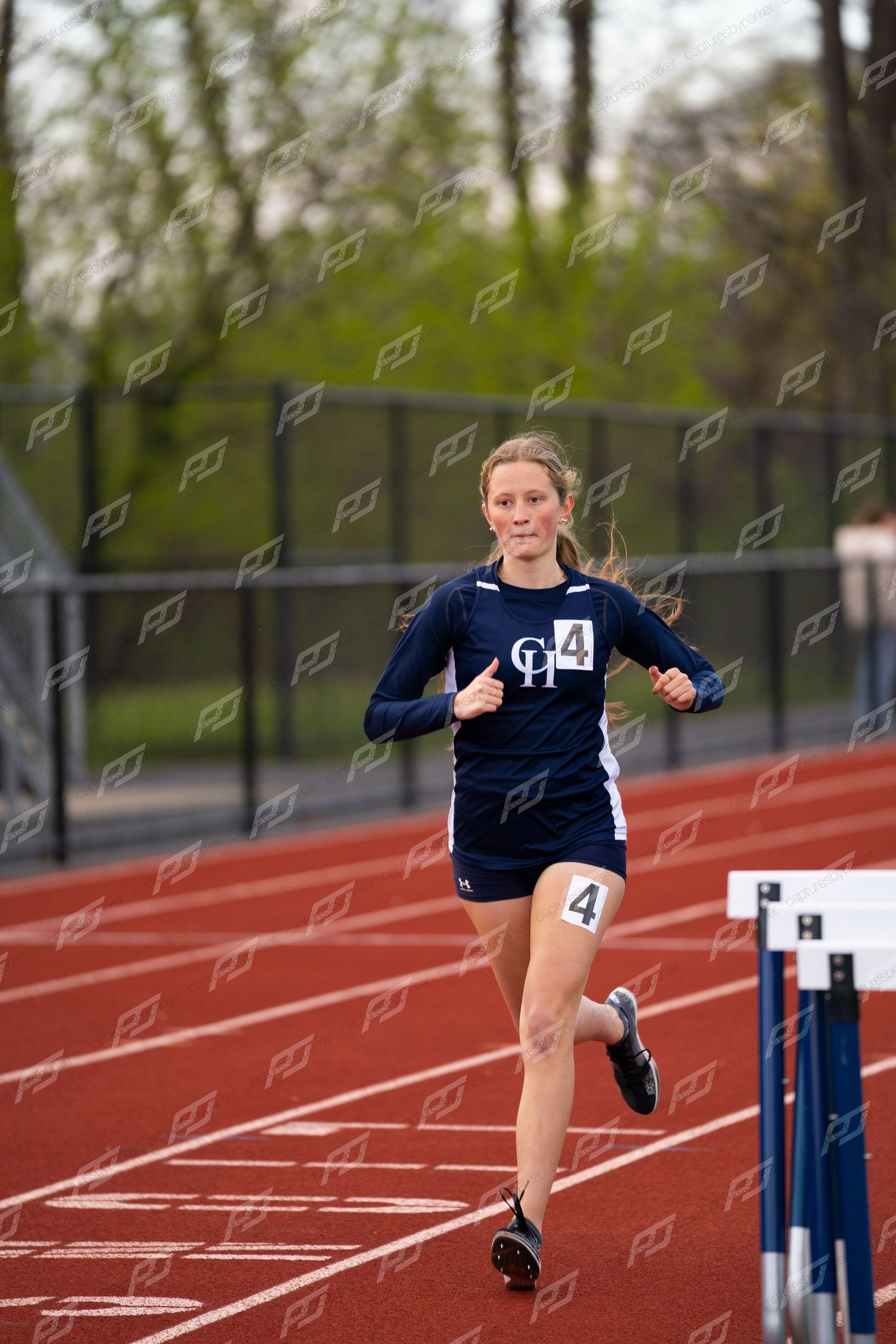 Female runner in navy uniform with number 4 competes on a red outdoor track near hurdles.