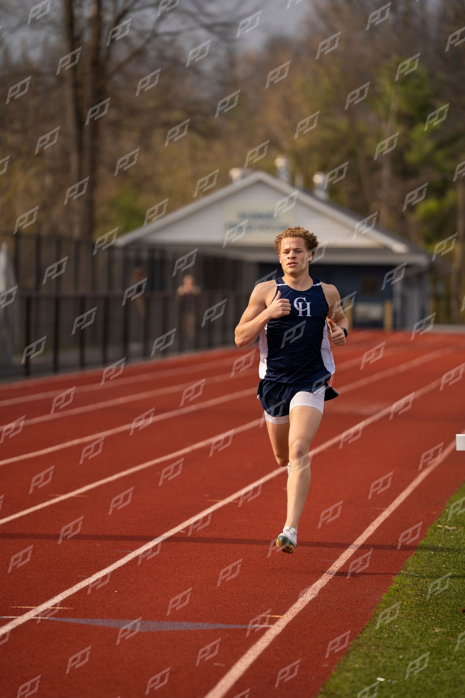 Male CH track runner in navy uniform sprinting on red track near building.