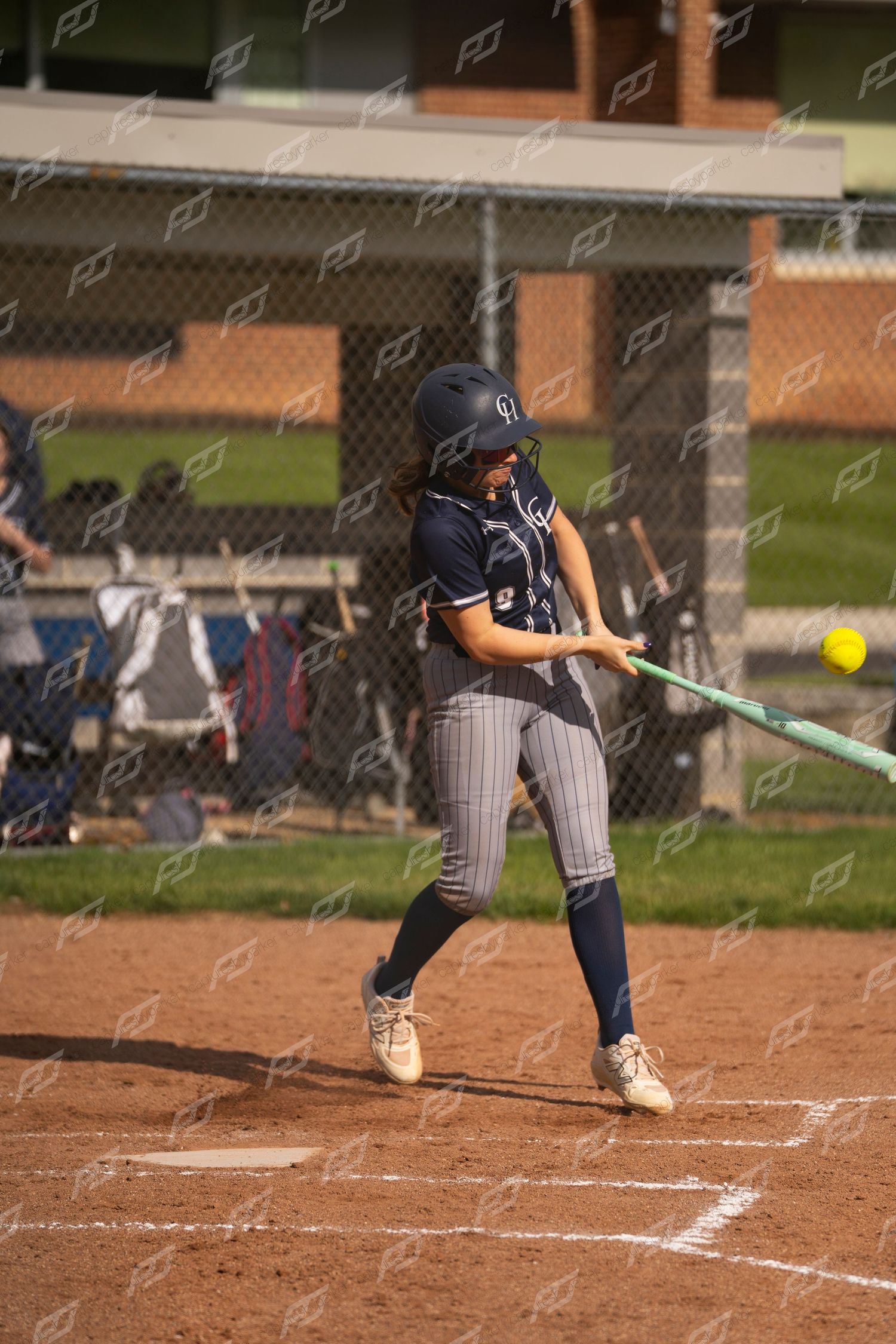 Female softball player in navy pinstripe uniform makes contact with a yellow softball at home plate.
