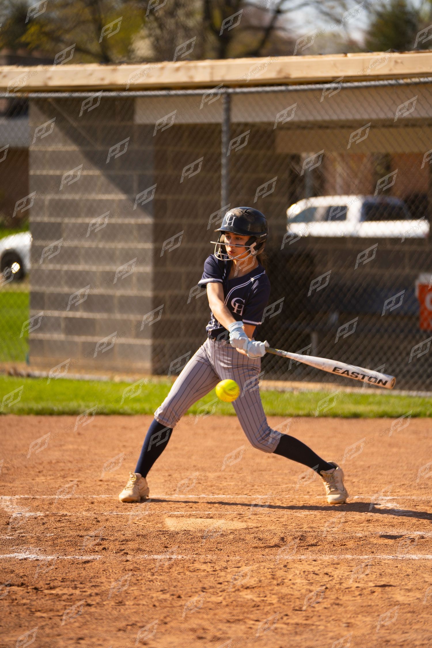 Softball batter in navy uniform swings at yellow ball with Easton bat during a game on a dirt field.
