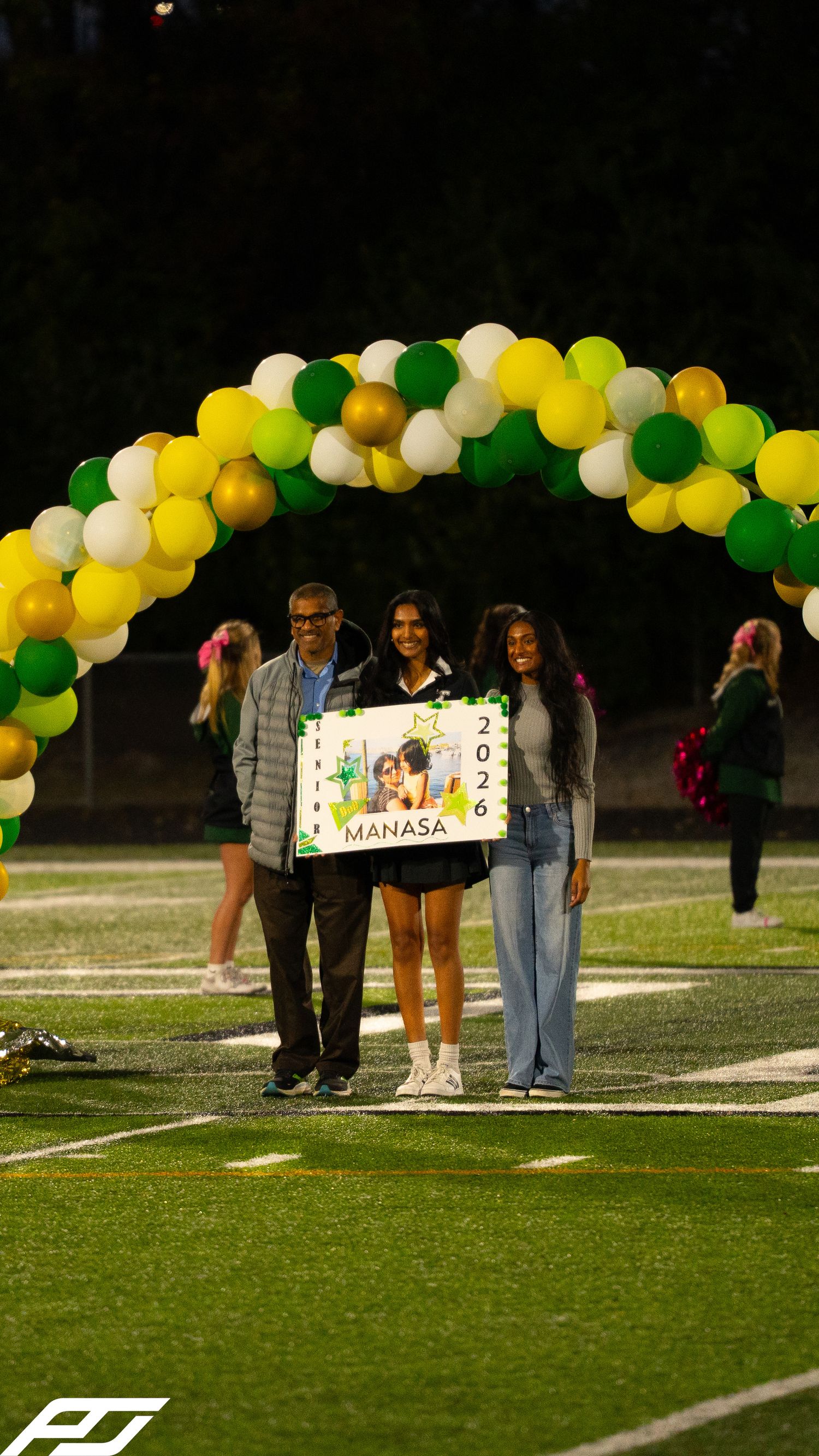 Manasa 2026 senior night celebration with family under colorful balloon arch on green football field at night.