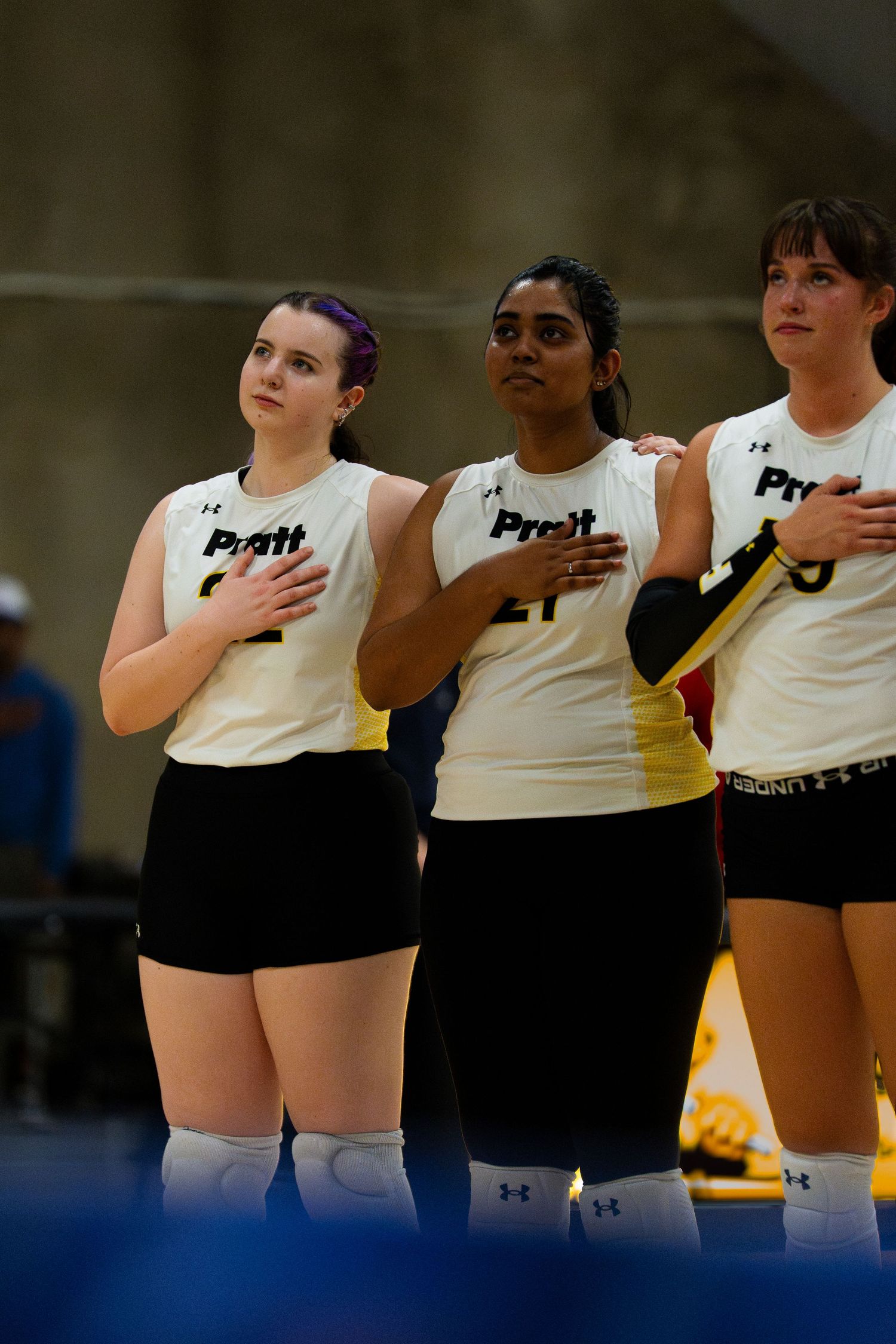Three Pratt volleyball players stand with hands on hearts during the national anthem in white uniforms.