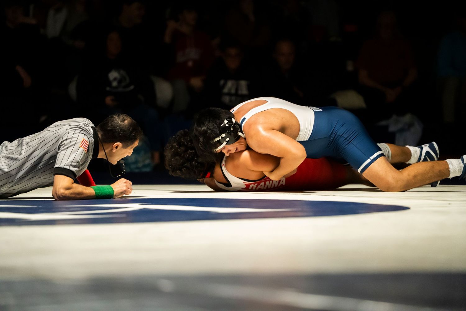 Referee watches closely as blue singlet wrestler pins opponent in red to the mat during wrestling match.