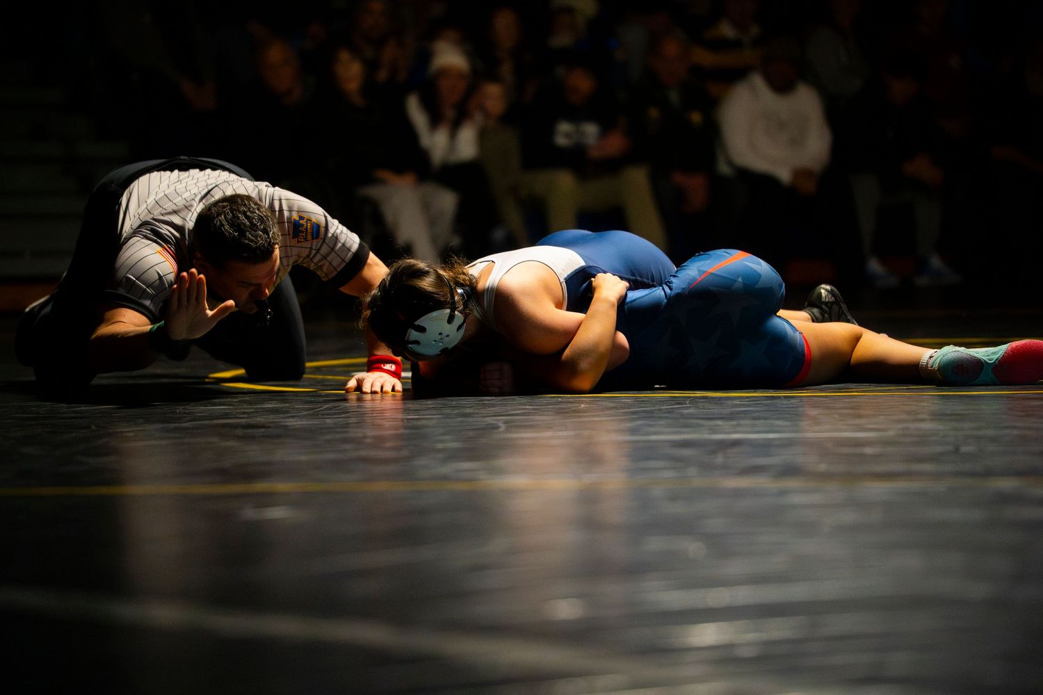 Two wrestlers compete on a mat during an indoor wrestling match under dramatic lighting.