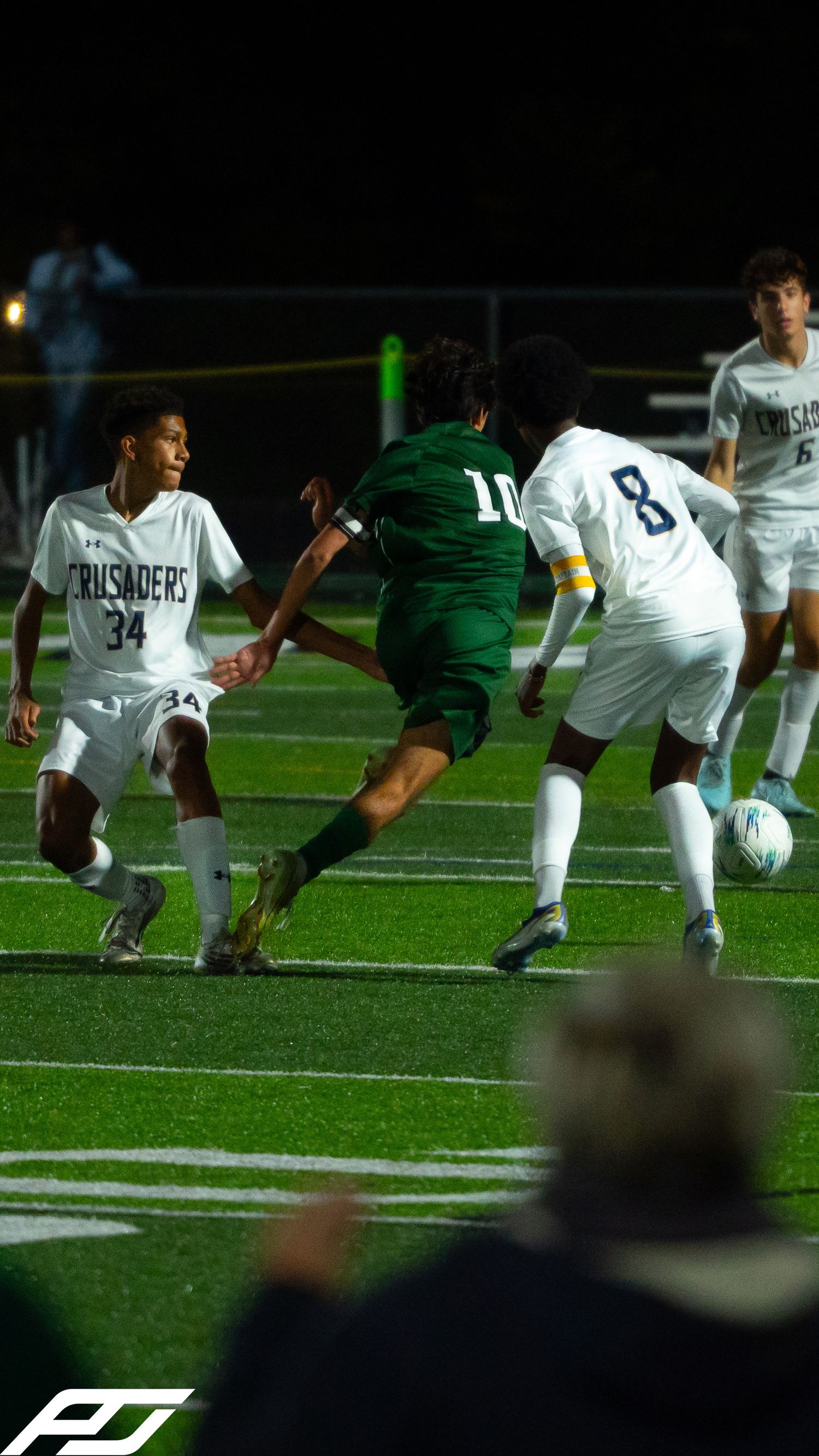 Player #10 in green dribbles between two Crusaders defenders #34 and #8 in white during a night soccer match.