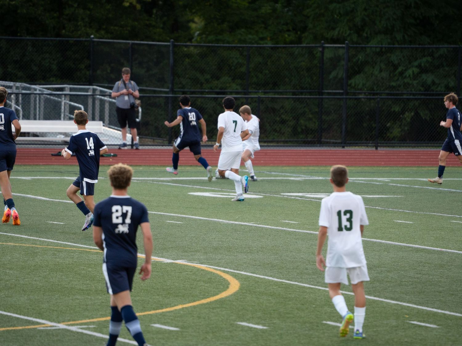 High school soccer players in white and navy uniforms chase ball on green turf field.