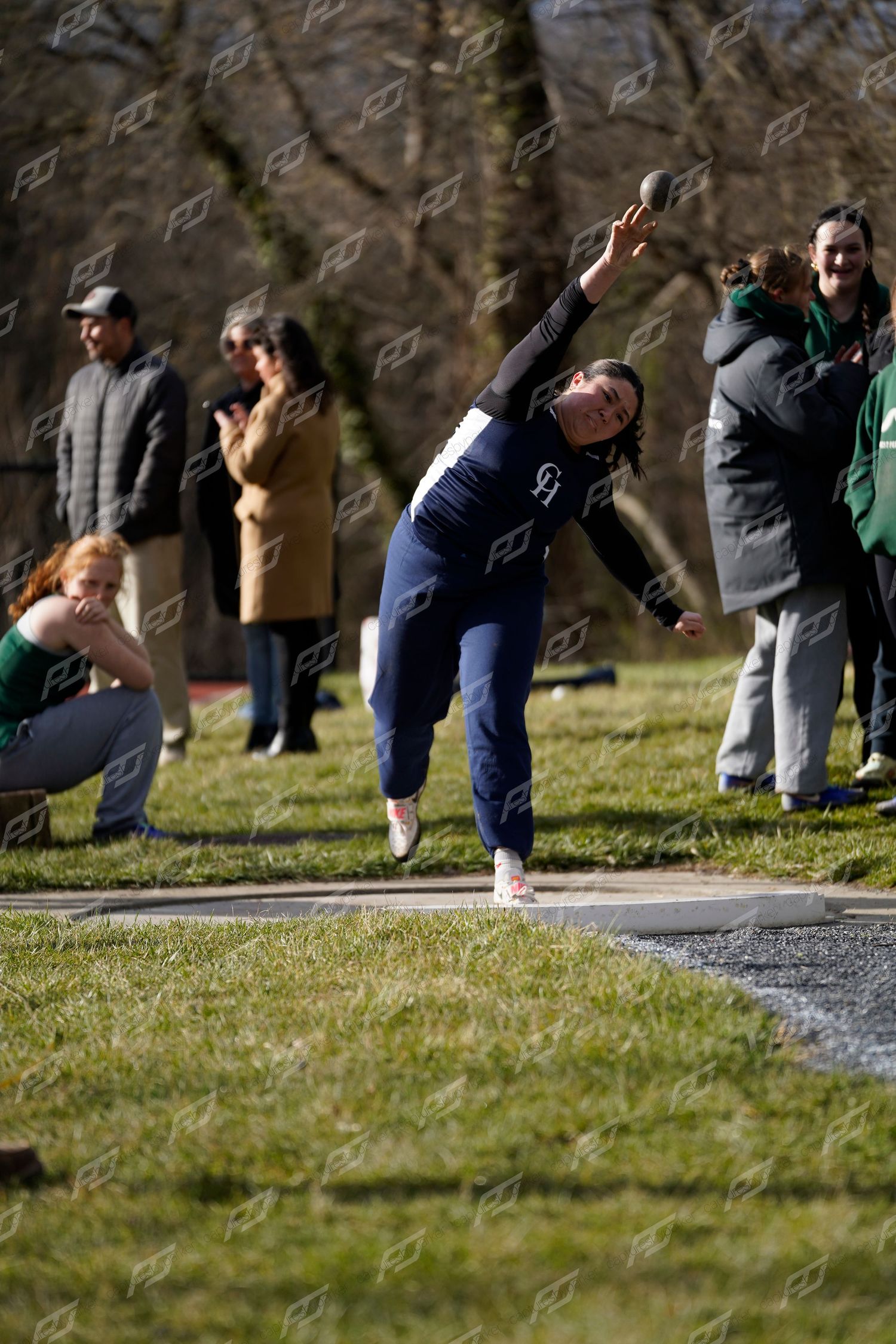 Female track athlete releasing shot put with full extension at an outdoor field competition.