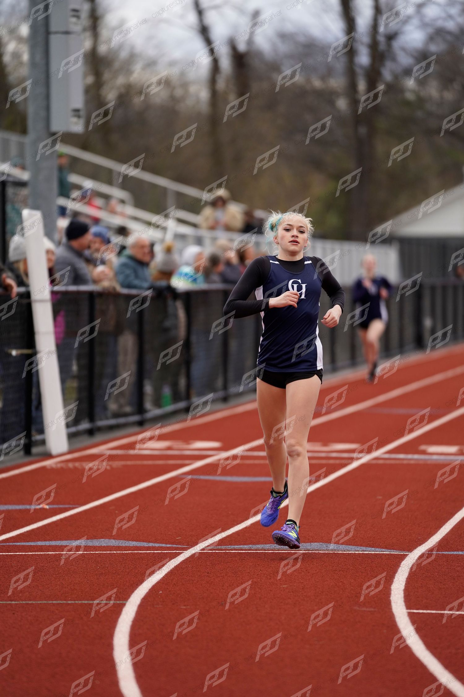 Female track athlete with blonde hair runs on a red outdoor track during a competitive meet.
