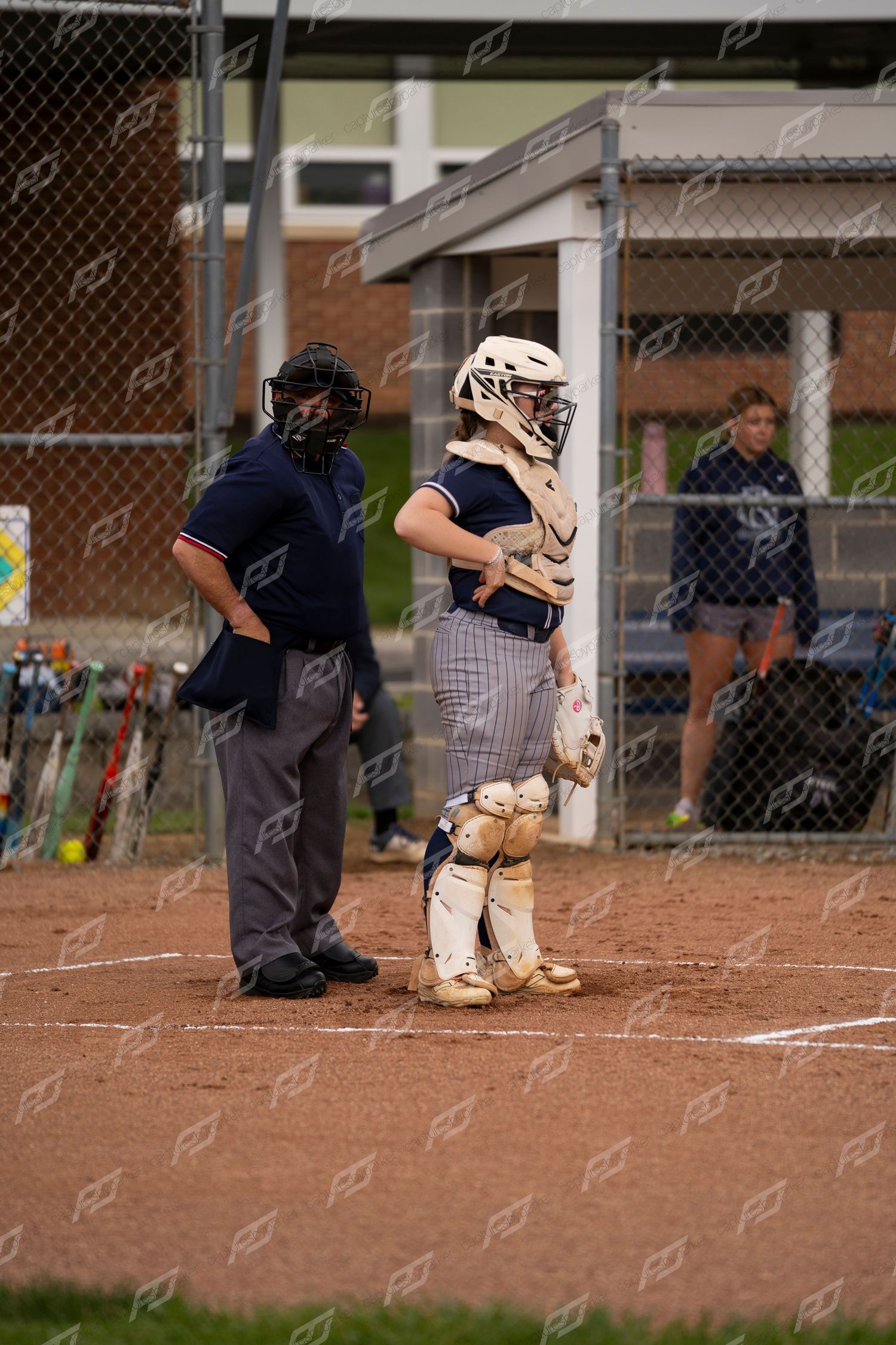 Softball catcher in full protective gear standing beside the home plate umpire between plays.