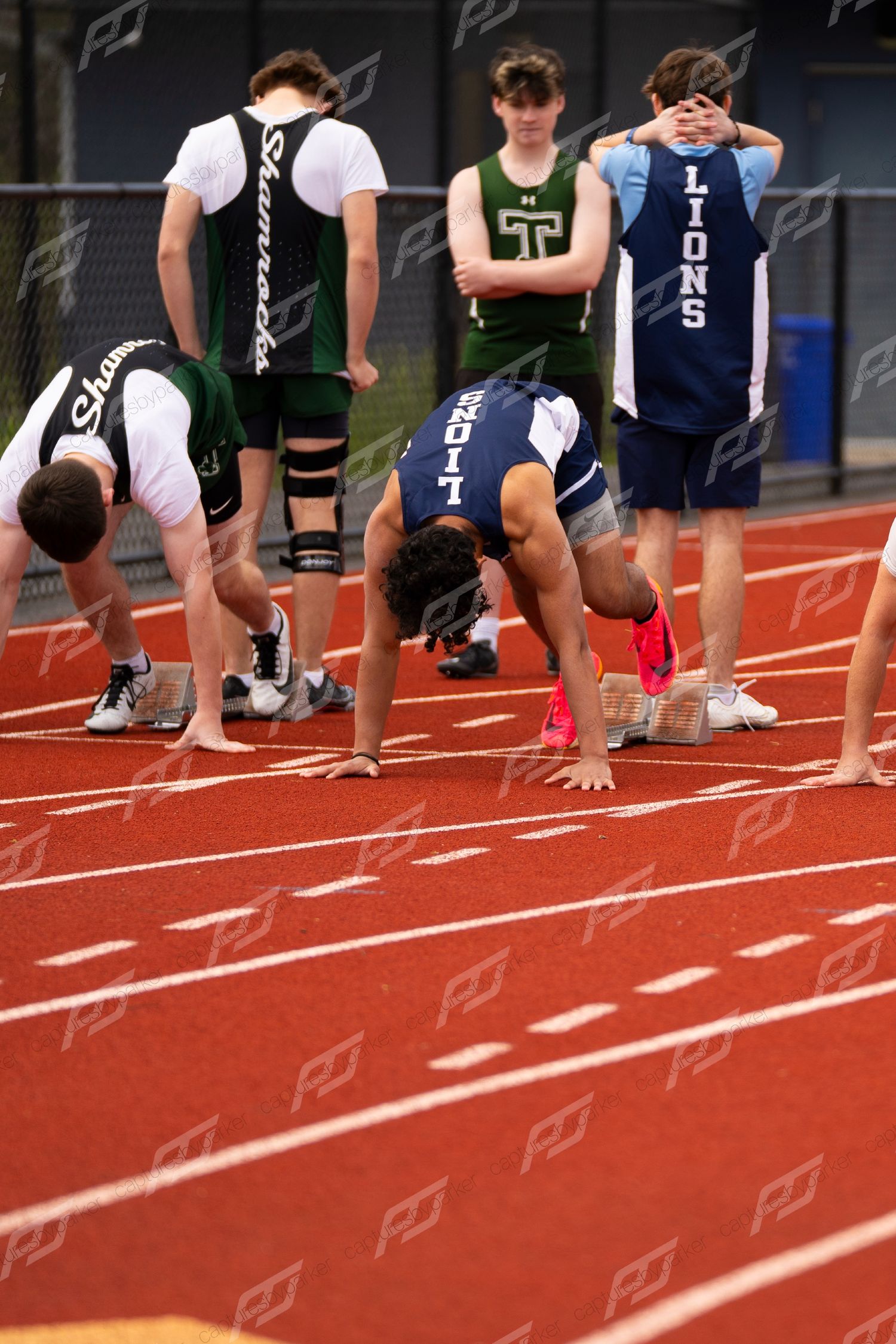 Male high school sprinters crouch at starting blocks on red track during outdoor track meet.