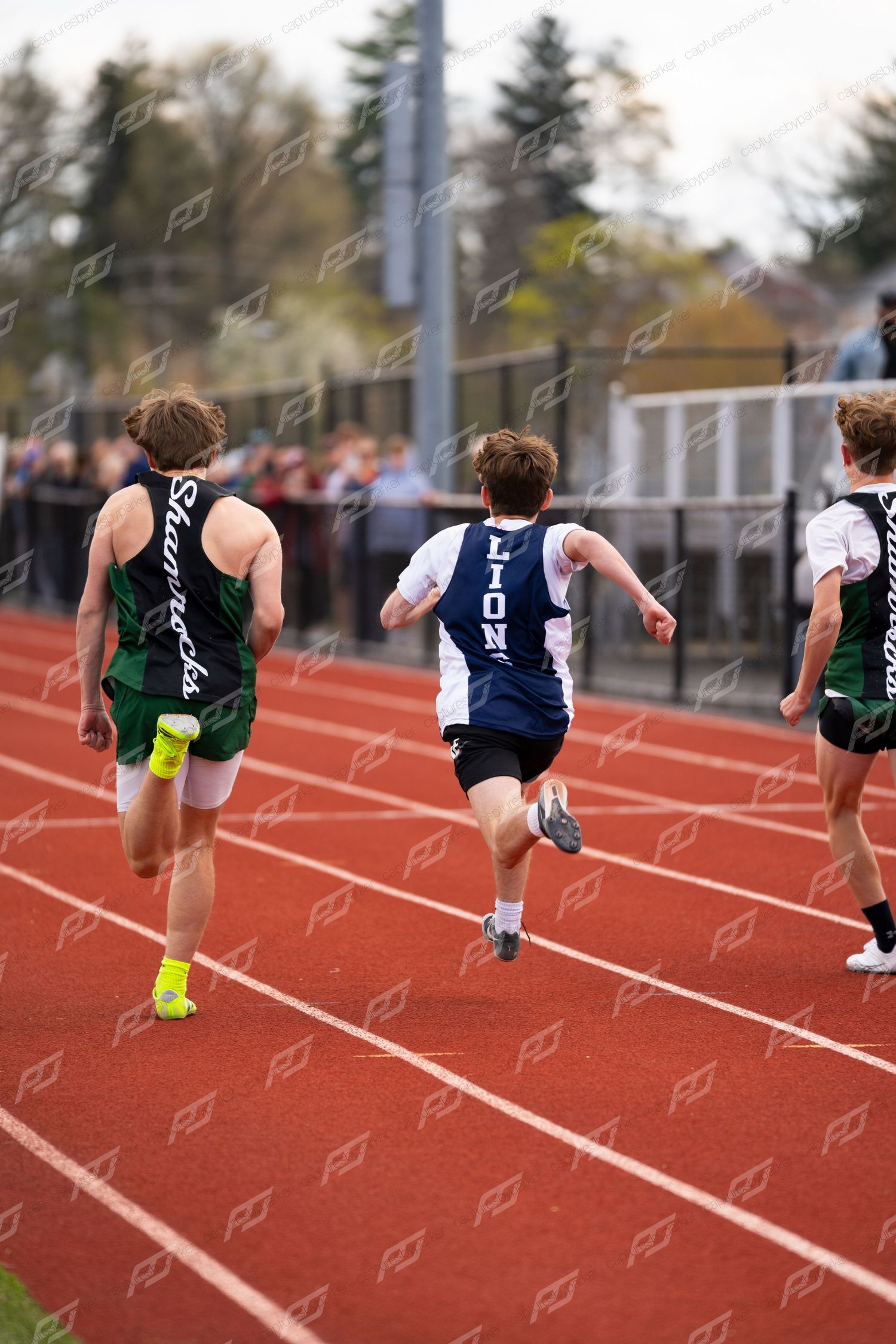 Three male runners in blue and green uniforms race down red track from behind during meet.