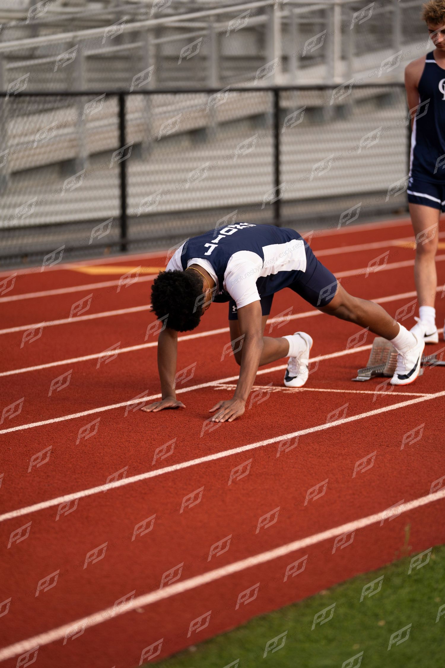 High school sprinter in navy uniform crouches at start position on red outdoor track.