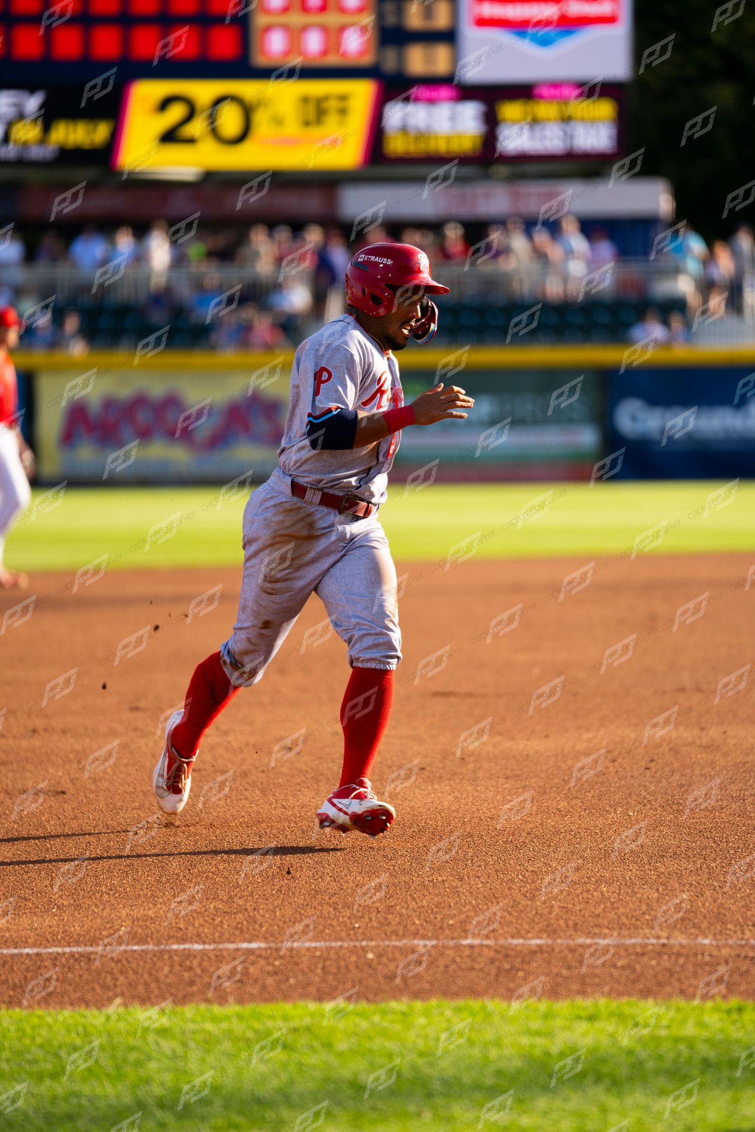 Baseball player in red uniform sprinting the bases during an evening game with scoreboard behind him.